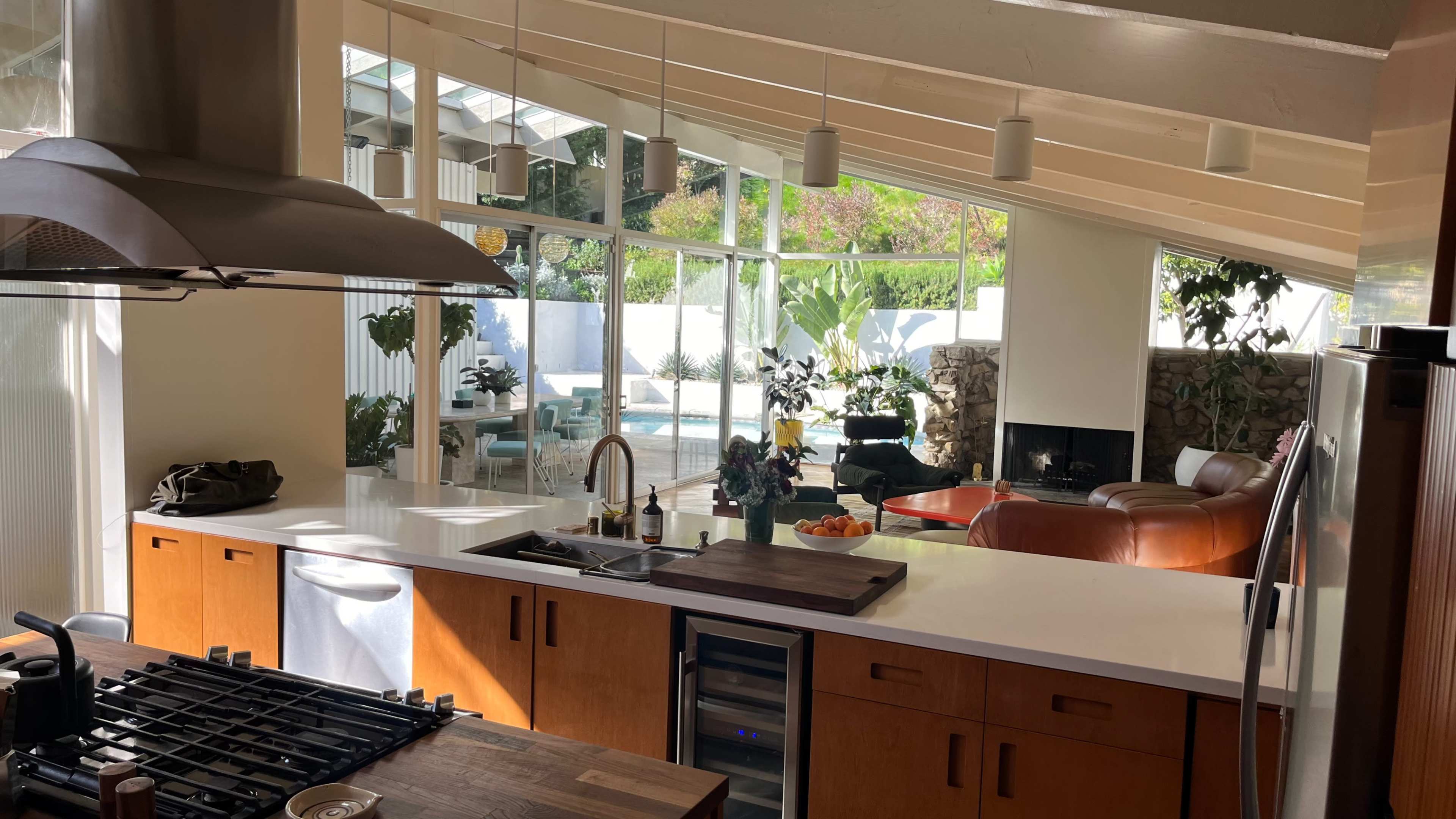 A spacious kitchen featuring a central island with a stovetop, adjacent to a living area filled with plants and a view of an outdoor pool through large windows.