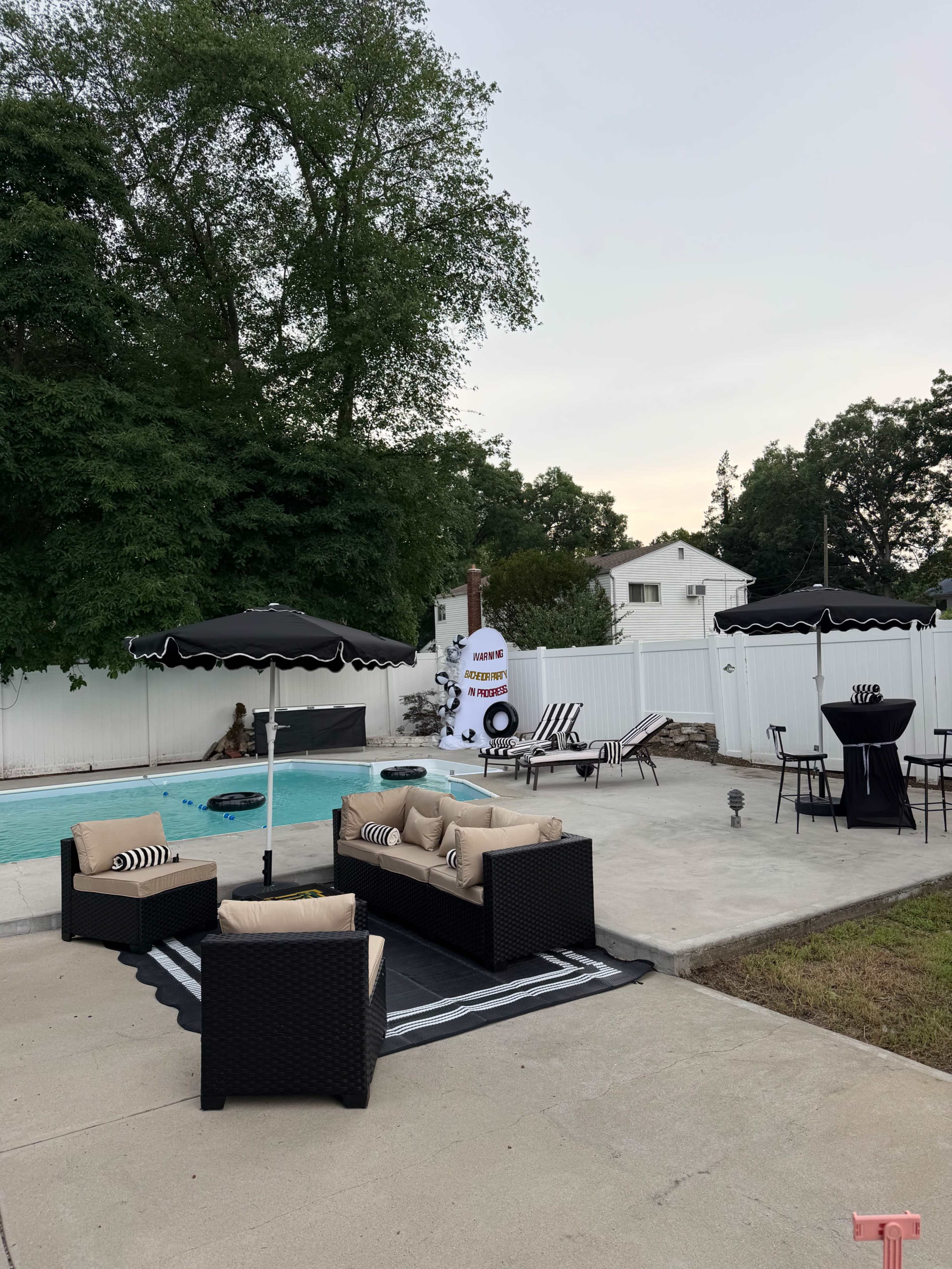 The scene features a backyard pool area with a seating arrangement of wicker sofas, a dining table, black and white umbrellas, and a decorative rocket structure in the background.