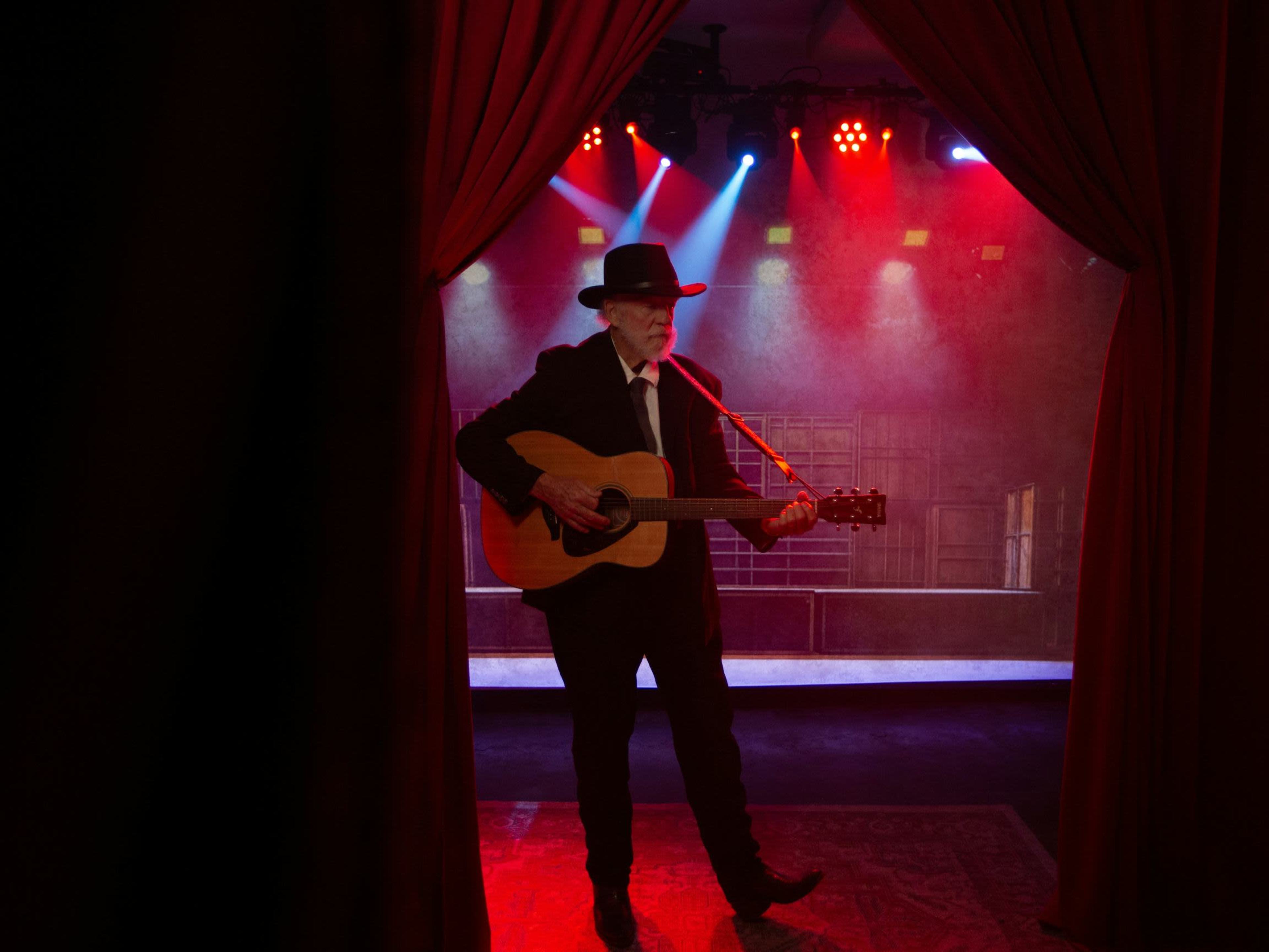 A man in a black suit and hat plays an acoustic guitar on stage, illuminated by colorful stage lights.