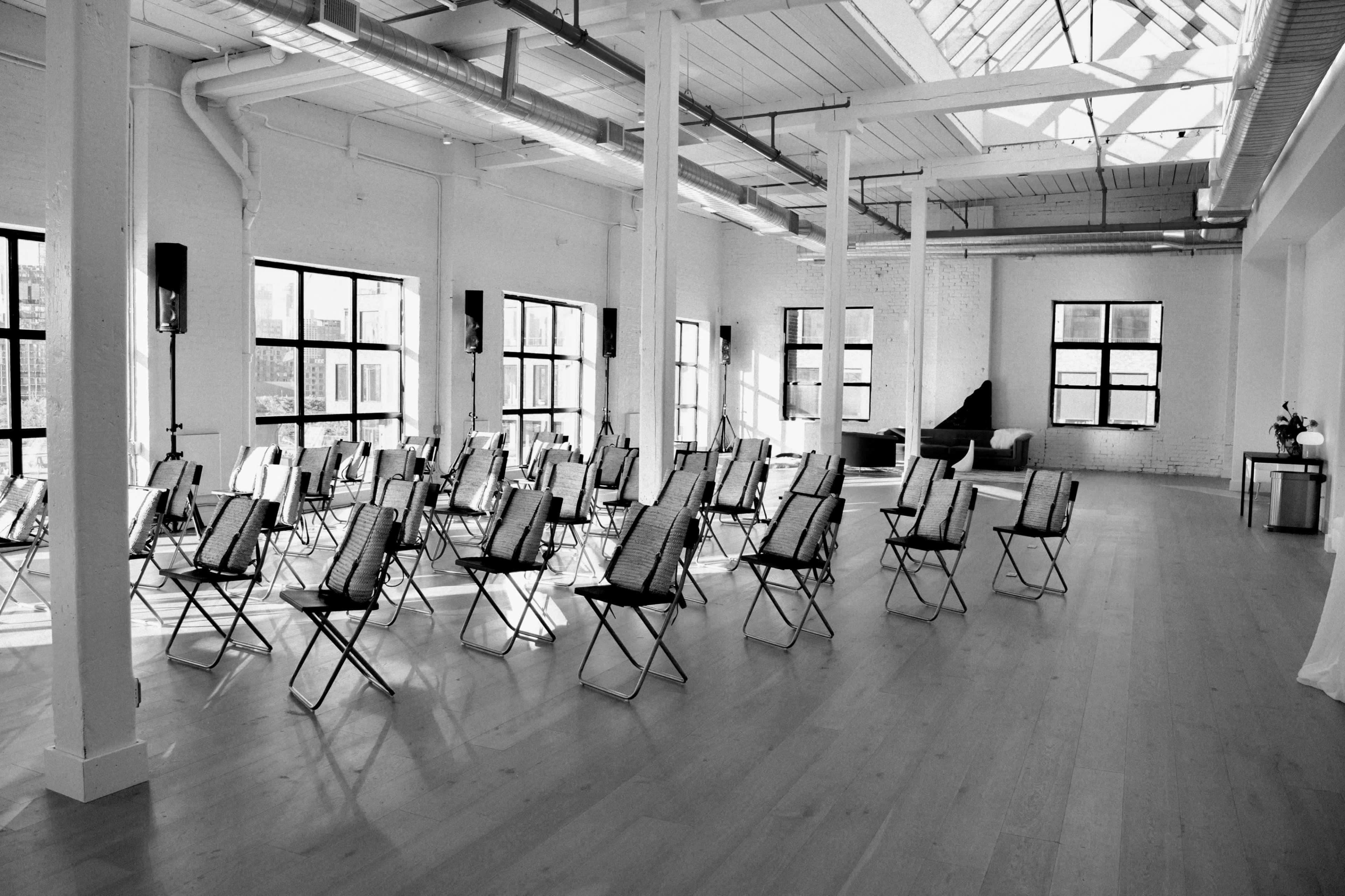A spacious, brightly lit room arranged with multiple rows of folding chairs facing a central area, with large windows providing natural light.