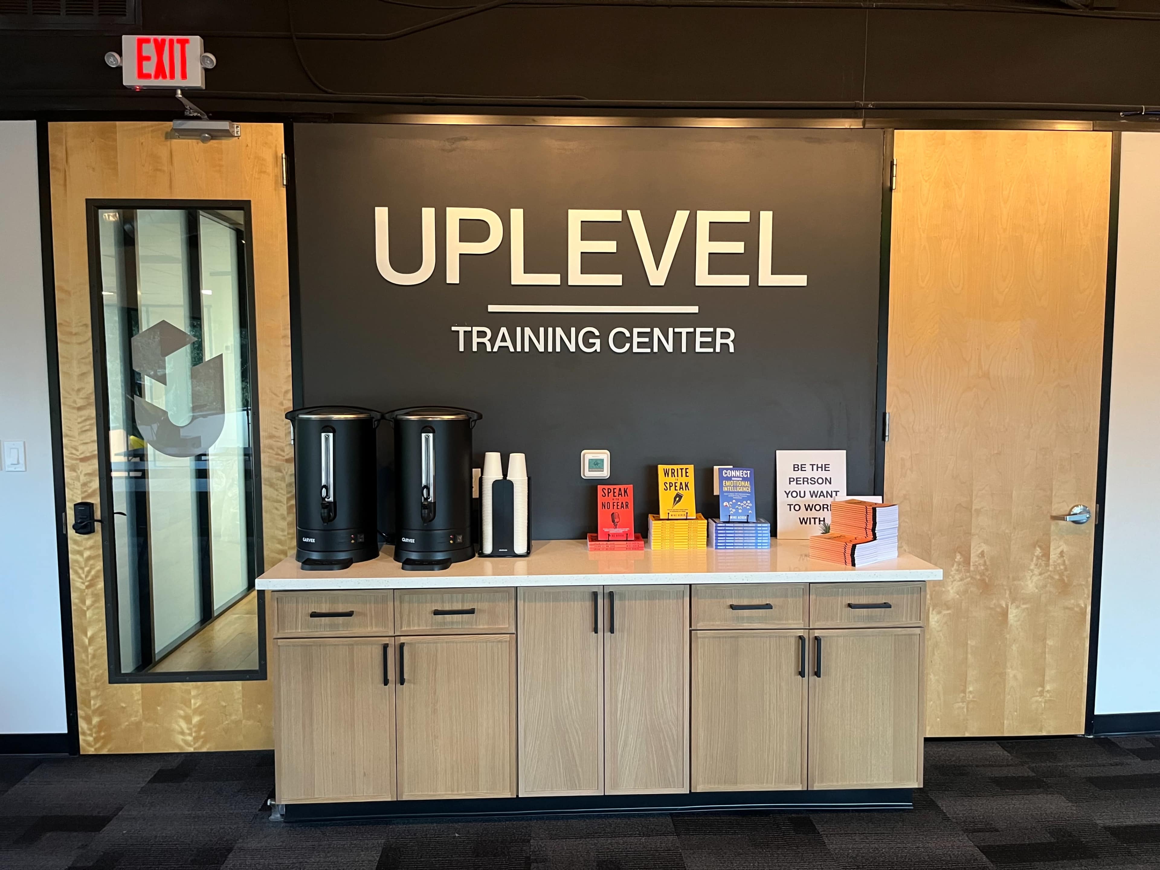 The image shows a training center with a wooden cabinet containing coffee dispensers, cups, and a shelf of books, with a prominent "UPLEVEL TRAINING CENTER" sign on the wall.