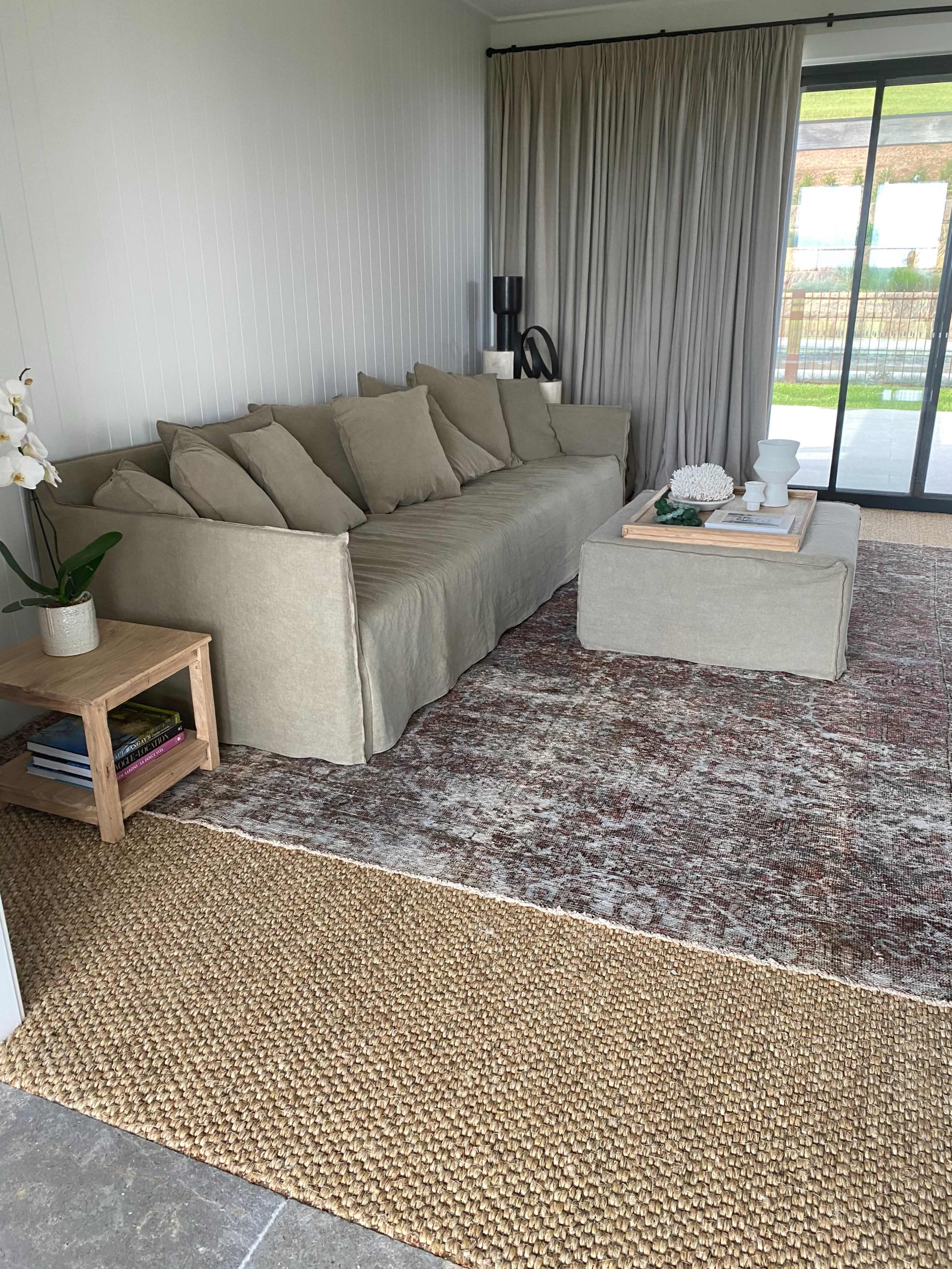 A modern living room featuring a beige sofa with numerous cushions, a wooden side table with books, a cream-colored rug, and large sliding glass doors leading to an outdoor area.