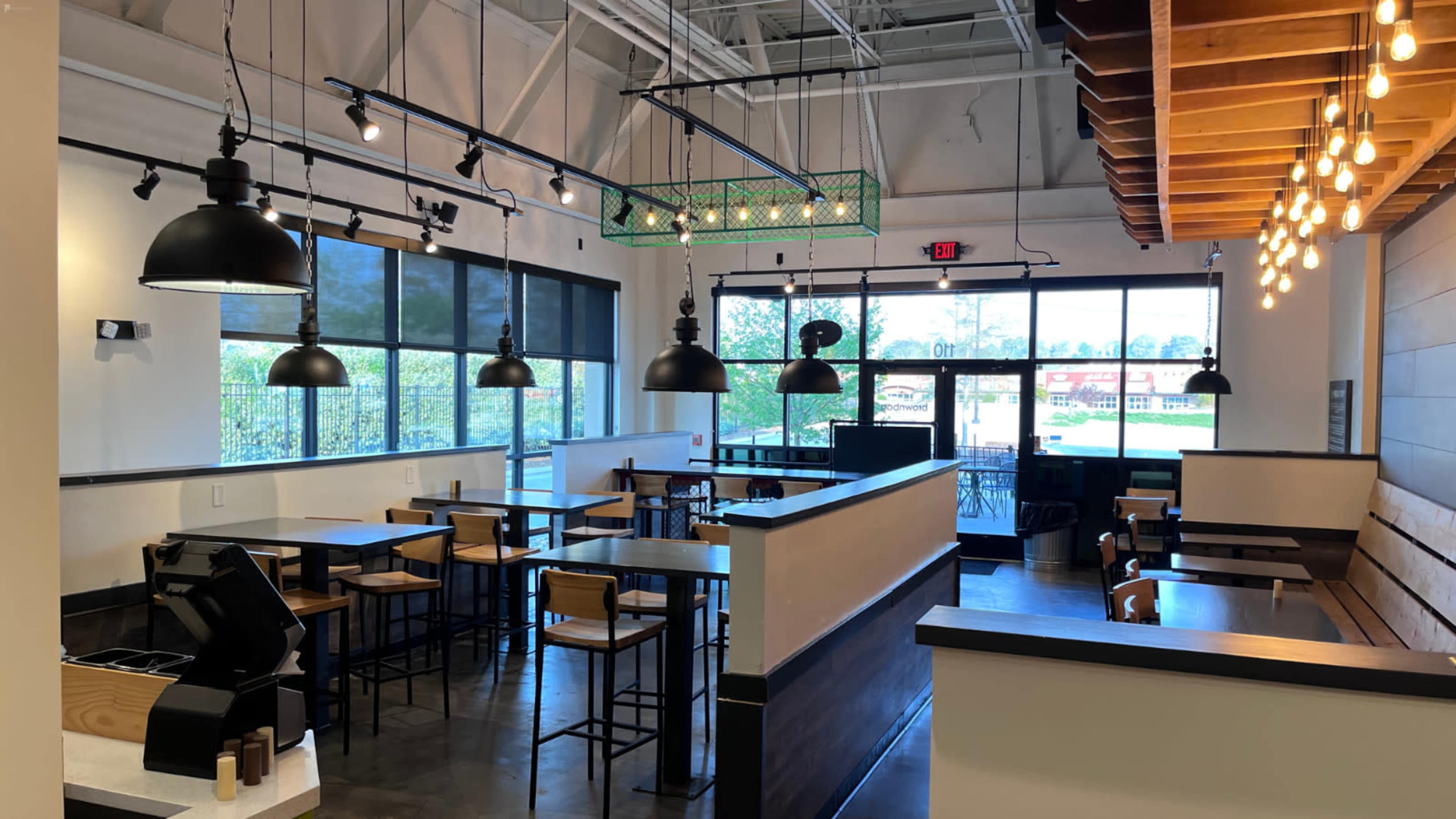 A modern cafe interior with black pendant lights, wooden tables and chairs, and large windows allowing natural light to enter.