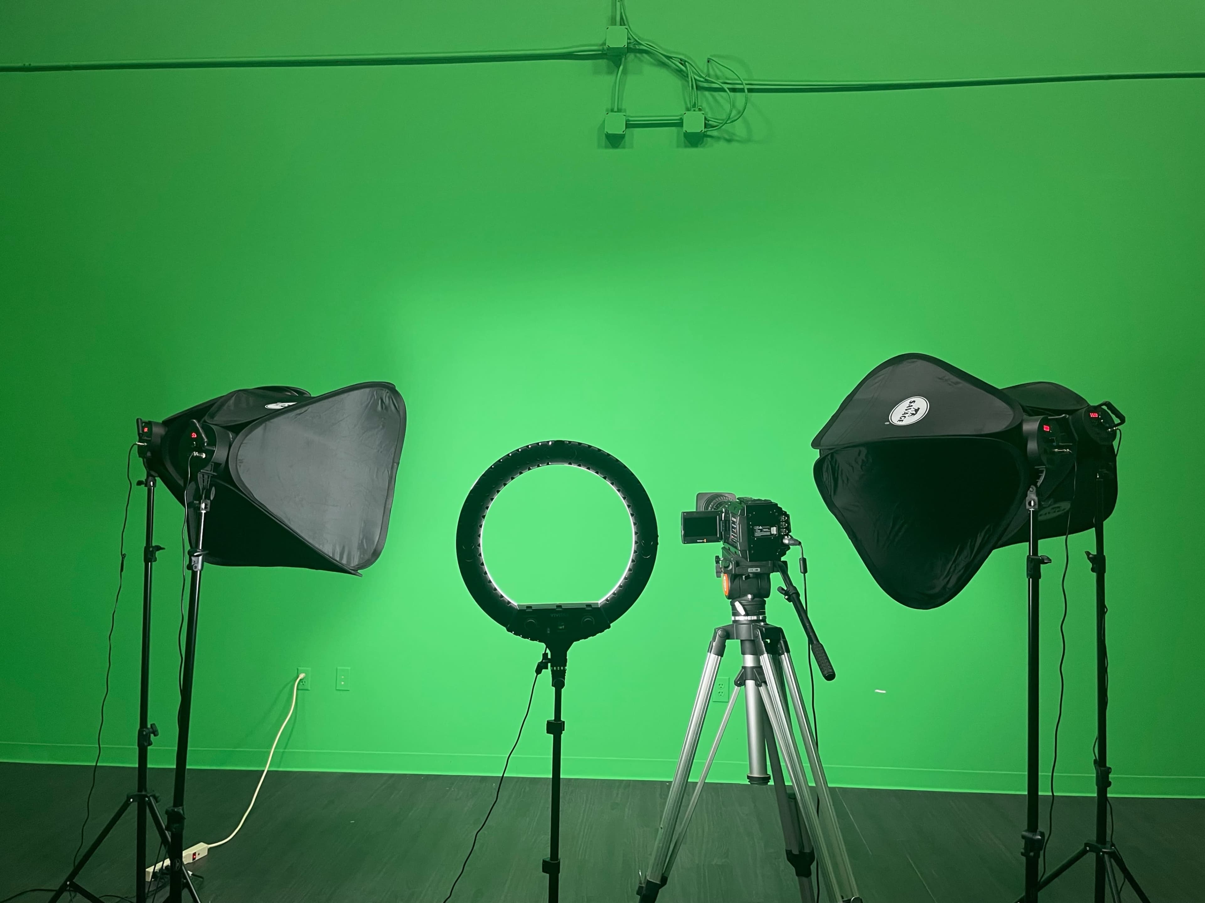A green screen studio setup with two softbox lights, a ring light, and a camera mounted on a tripod.