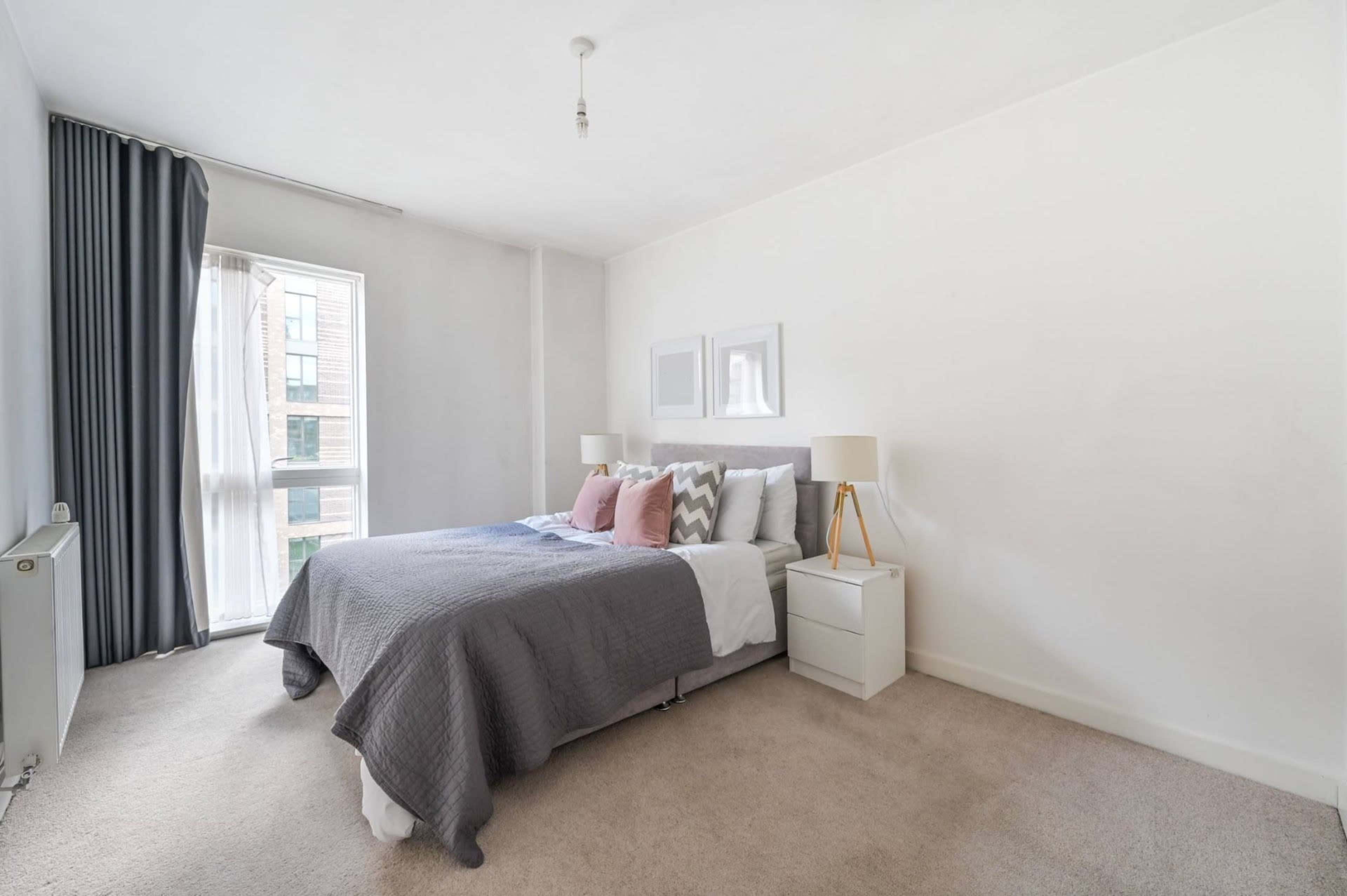 A neatly organized bedroom features a bed with gray bedding, two lamps on bedside tables, and large windows with light-colored curtains.