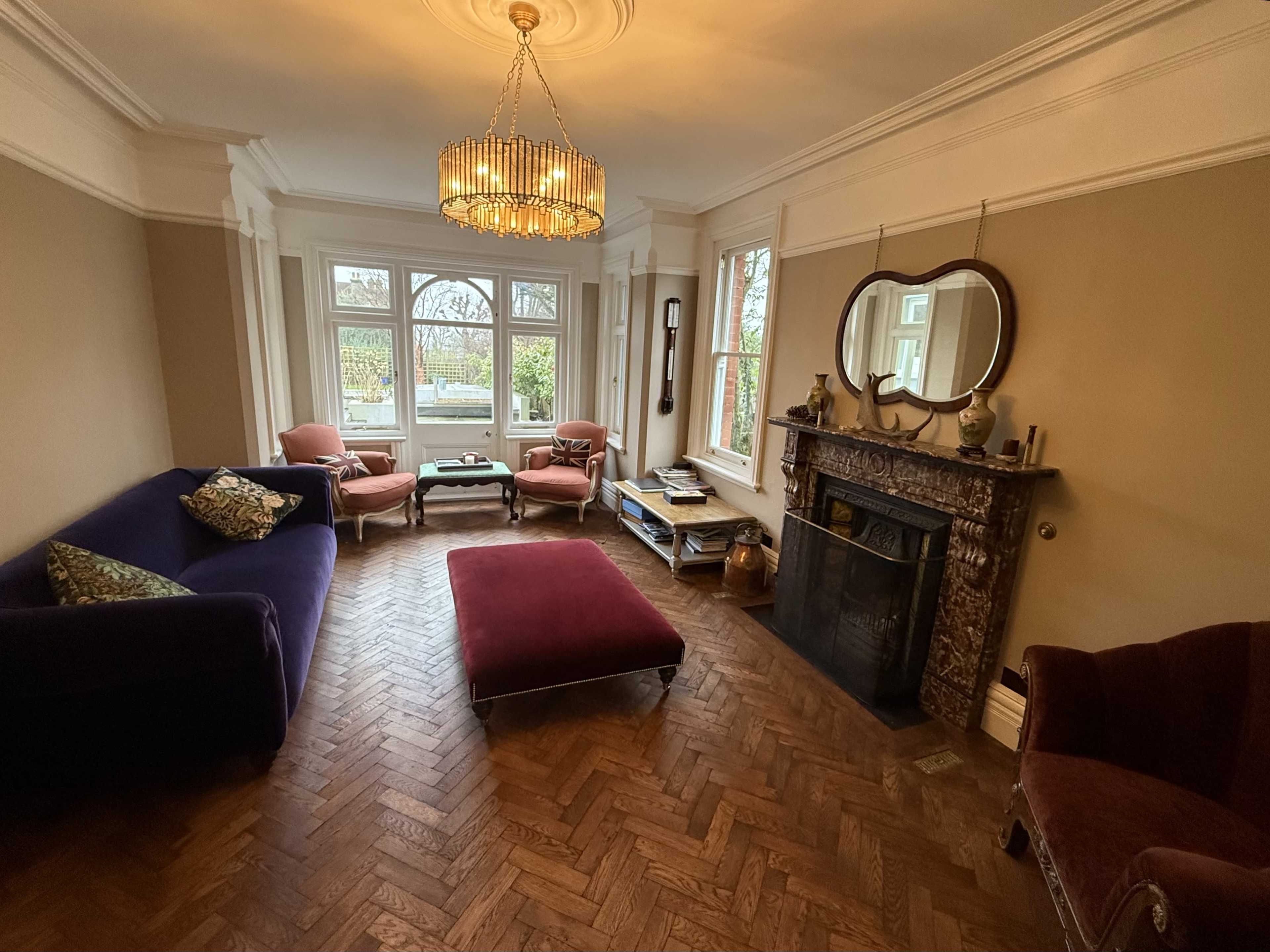 The image shows a spacious living room with a chandelier, a purple sofa, and a marbled fireplace, featuring large windows that provide natural light.