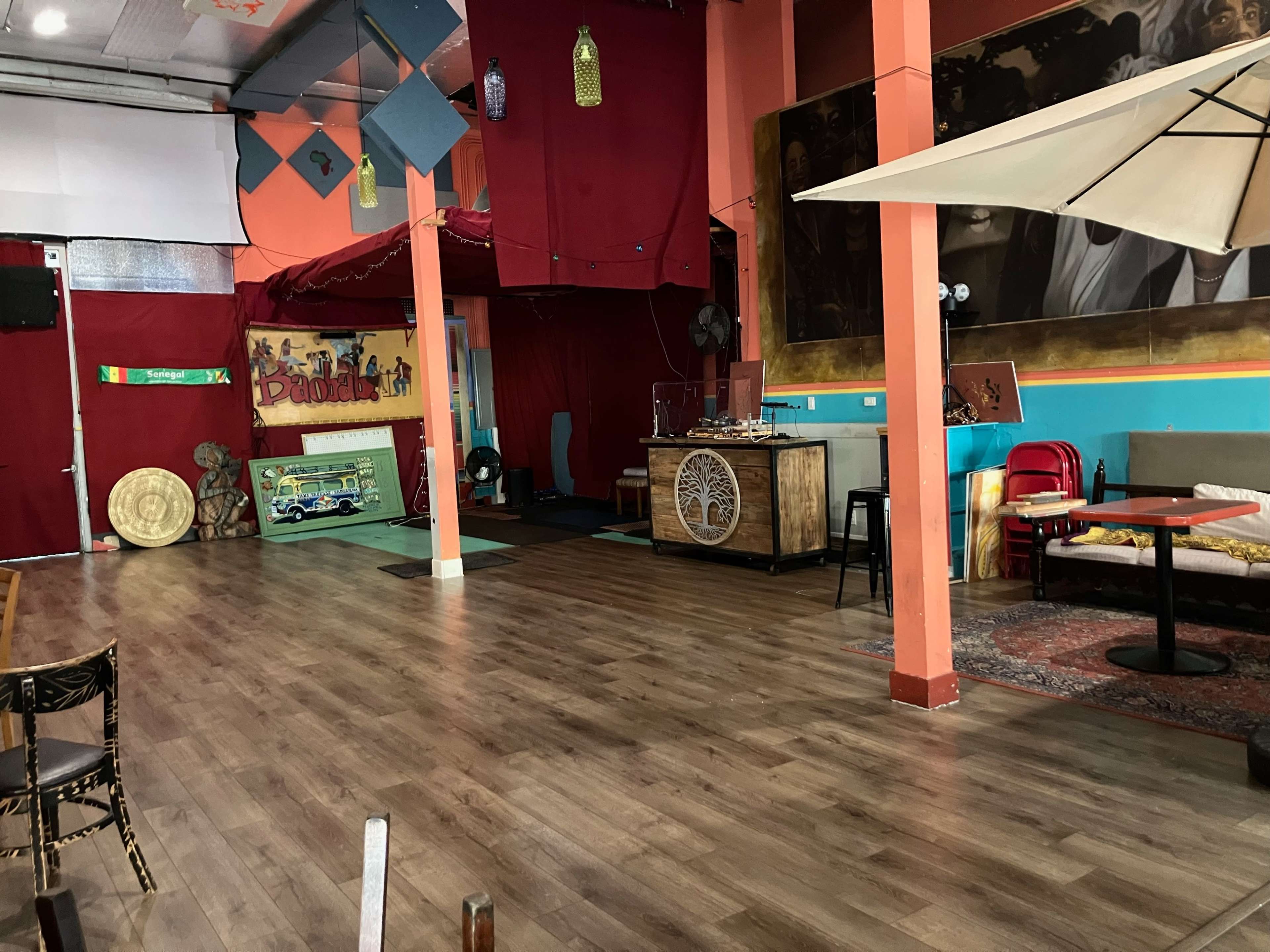 The image shows a spacious, brightly colored room with wooden flooring, scattered furniture, and decorative elements like fans and artwork on the walls.
