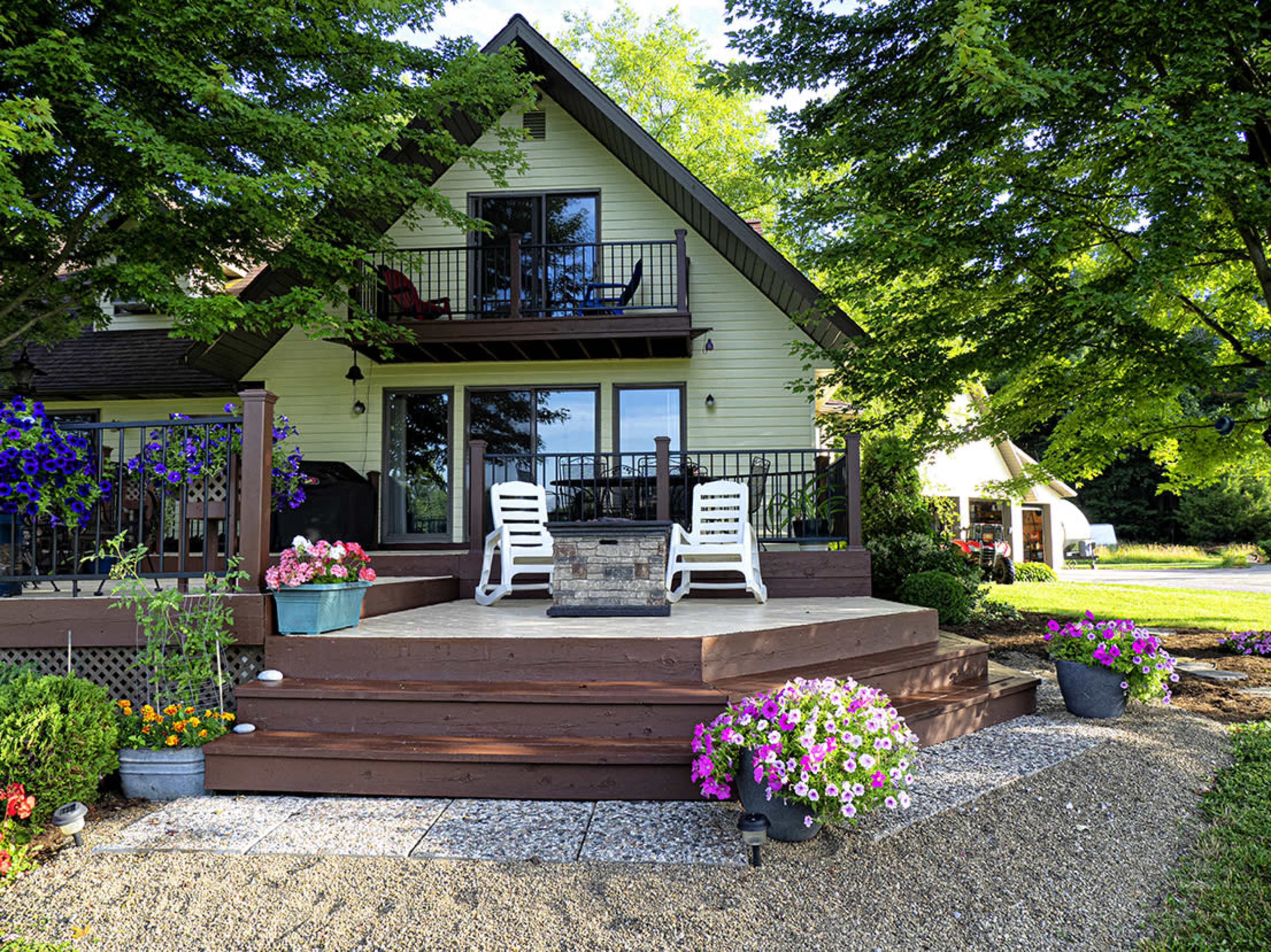 A two-story house with a sloped roof and a spacious wooden deck is surrounded by colorful flowering plants and lush green trees.
