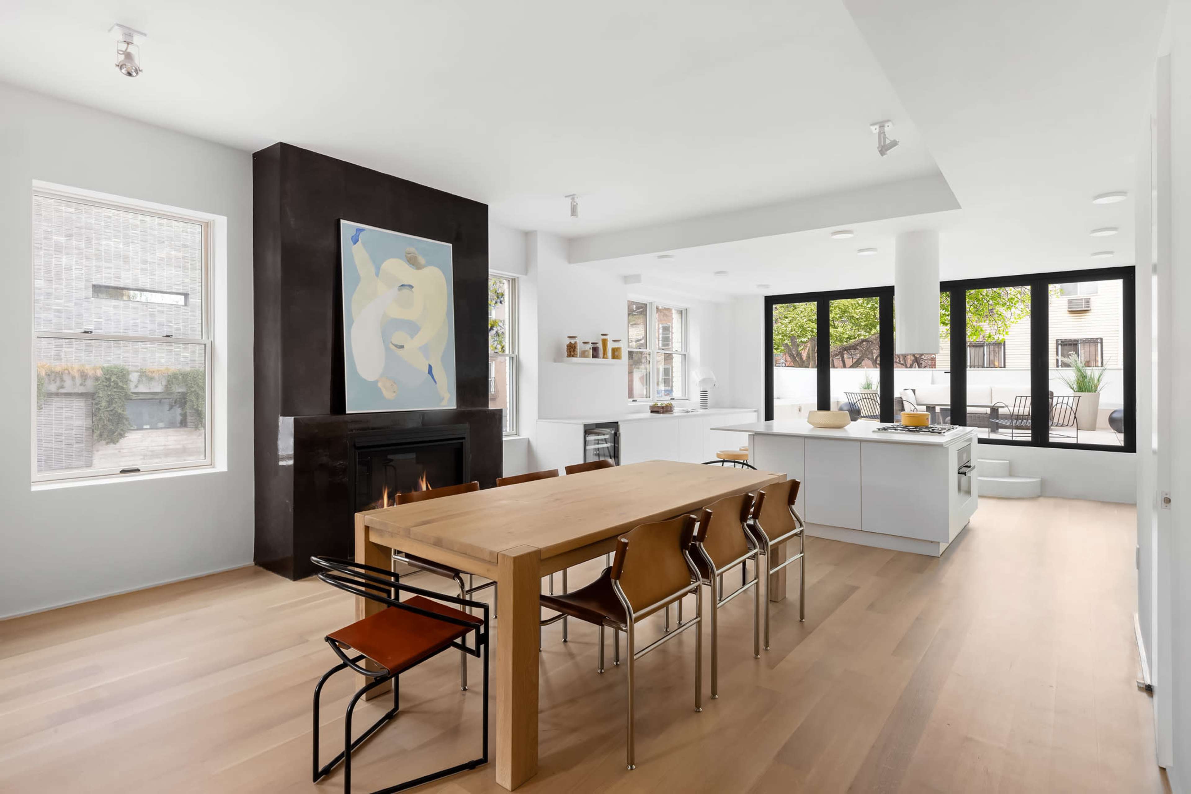 The image shows a modern kitchen and dining area with a large wooden table surrounded by chairs, a minimalist design, and large windows allowing natural light.