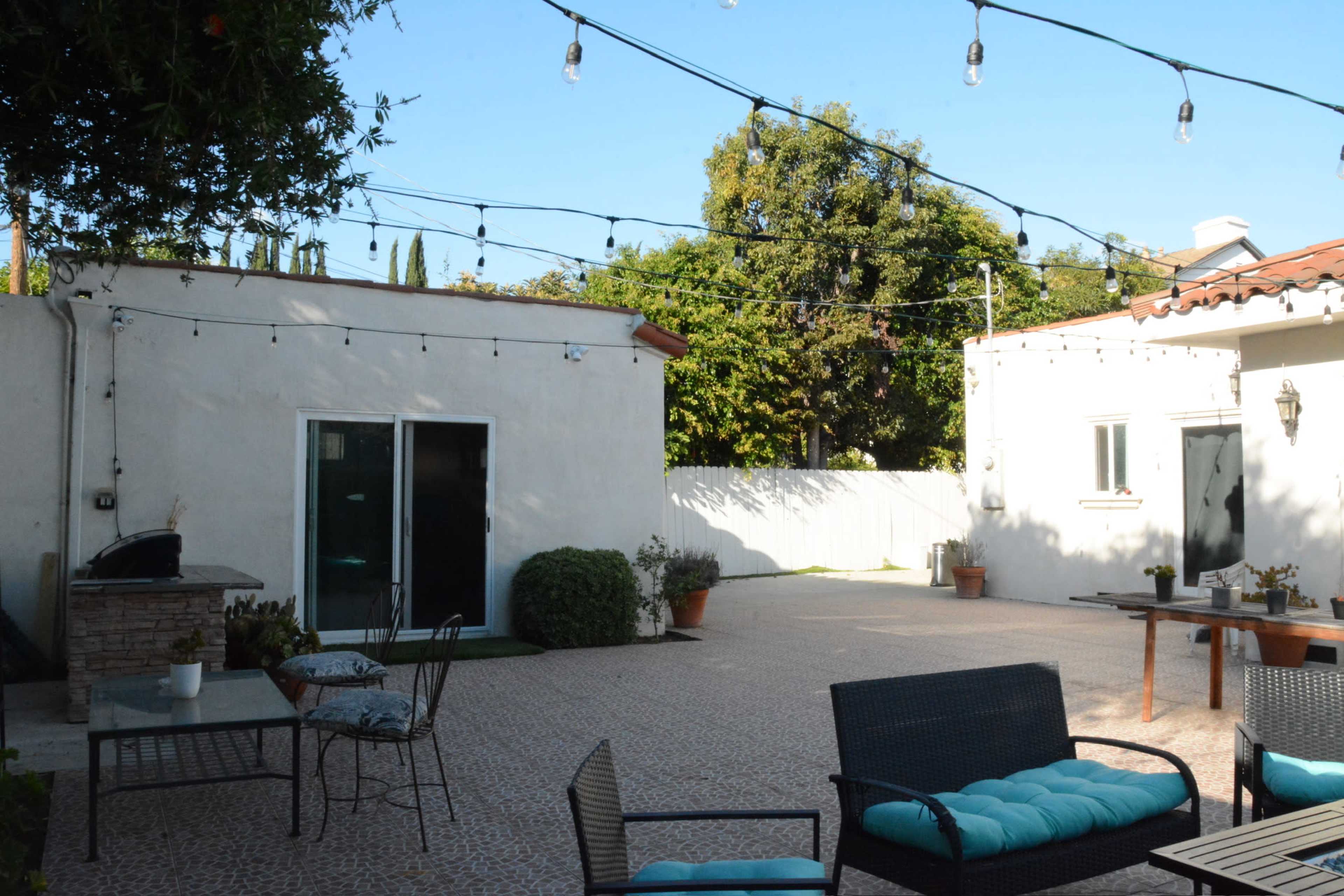 A spacious outdoor patio features string lights overhead, a seating area with dark wicker furniture and blue cushions, and a grill near the entrance of a white building.