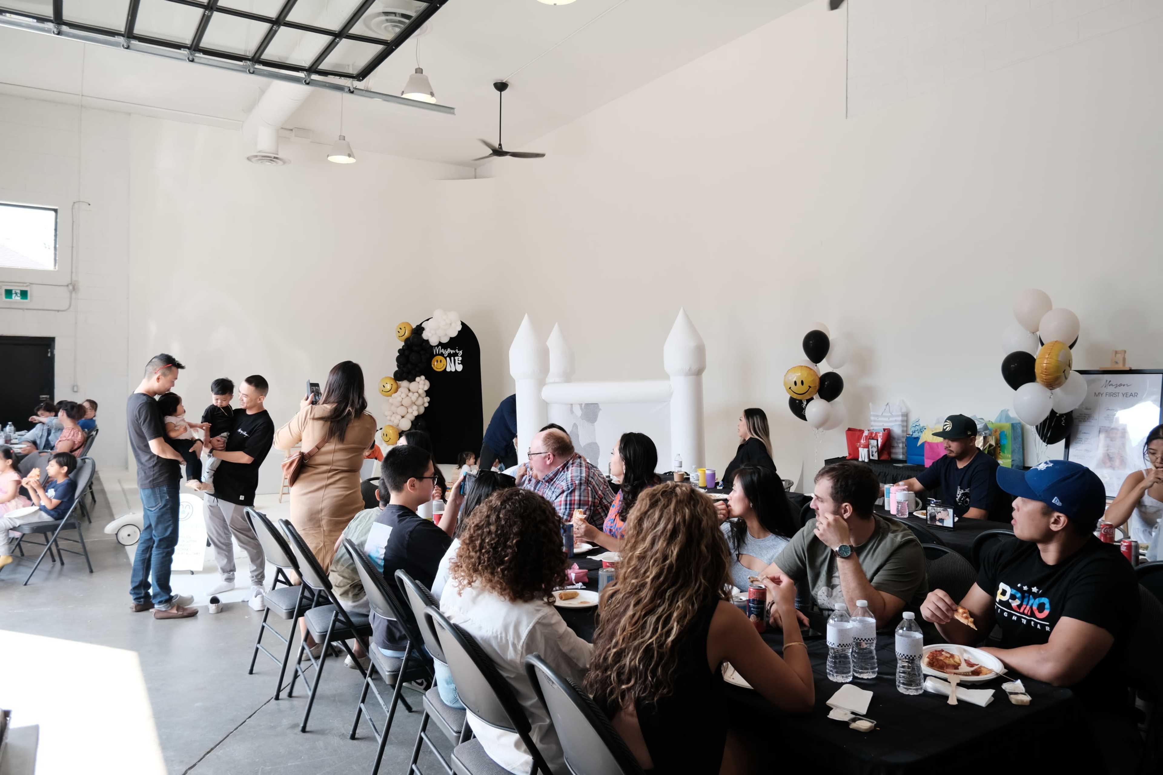 A group of people gather around tables in a spacious room decorated for a celebration, with balloons and a thematic backdrop.