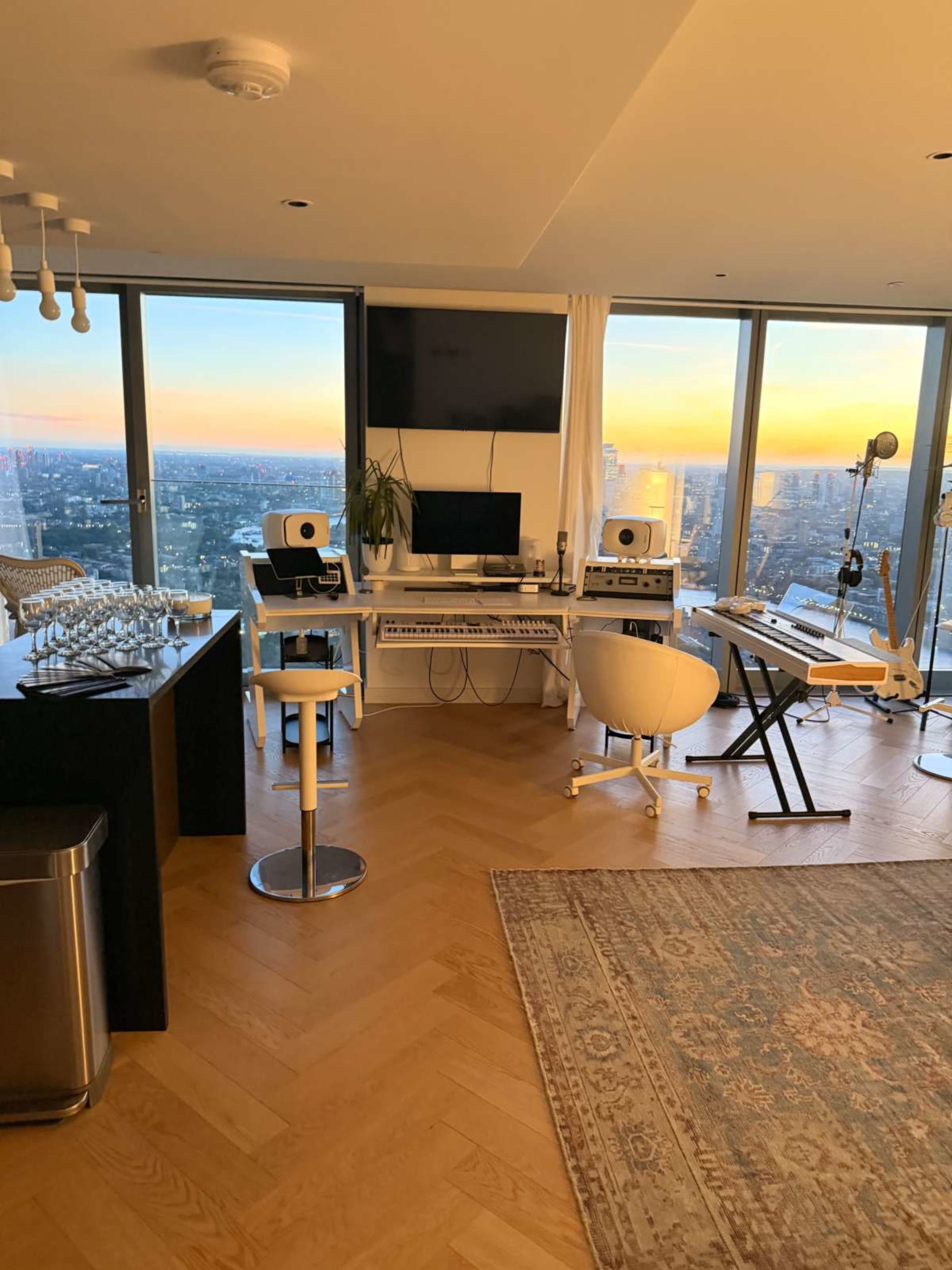 The image shows a modern room with a music production setup featuring a keyboard, monitor, and audio equipment, alongside large windows offering a view of a city skyline at sunset.