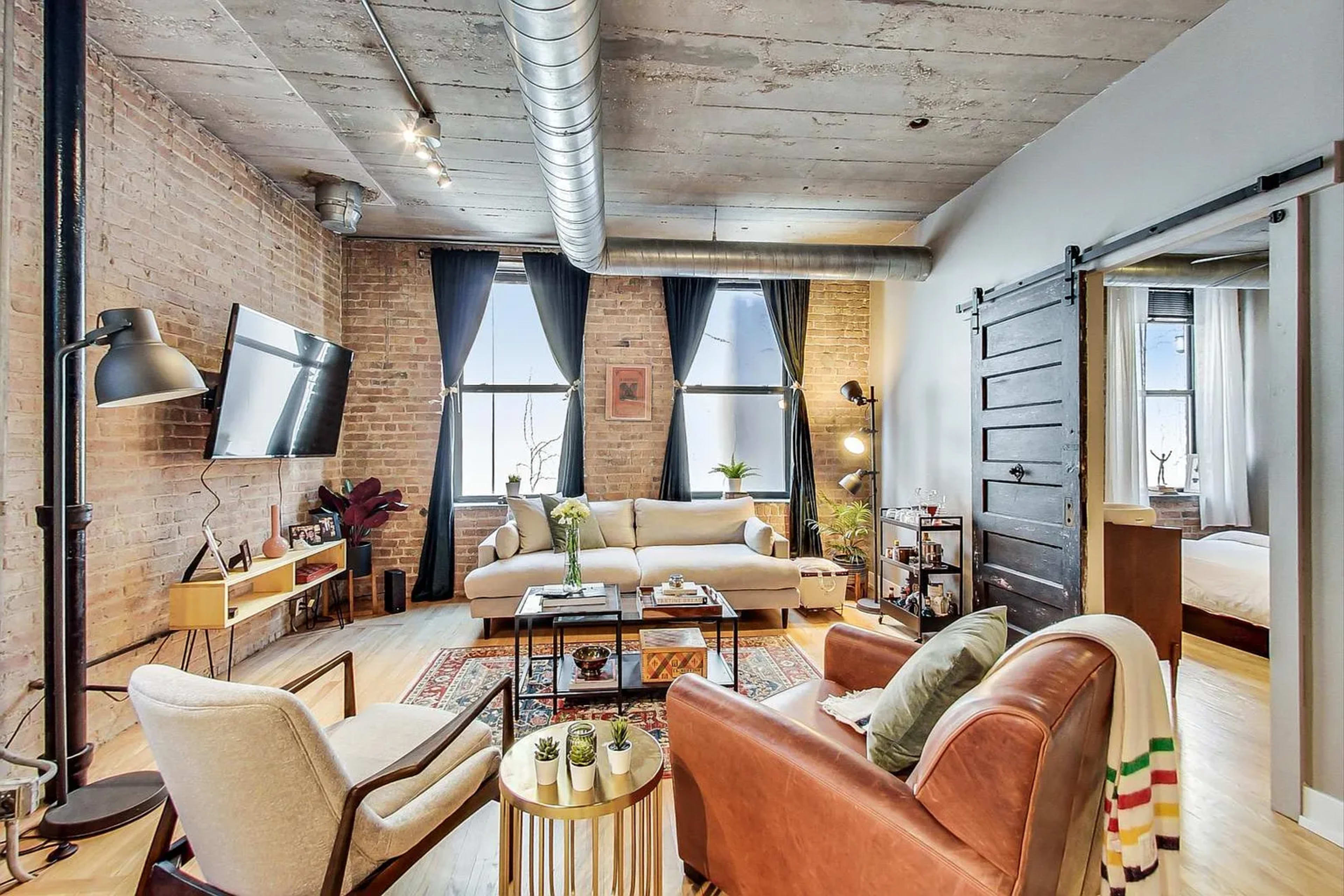 The image shows a modern living room featuring a mix of comfortable seating, a television mounted on a brick wall, and large windows allowing natural light to enter the space.