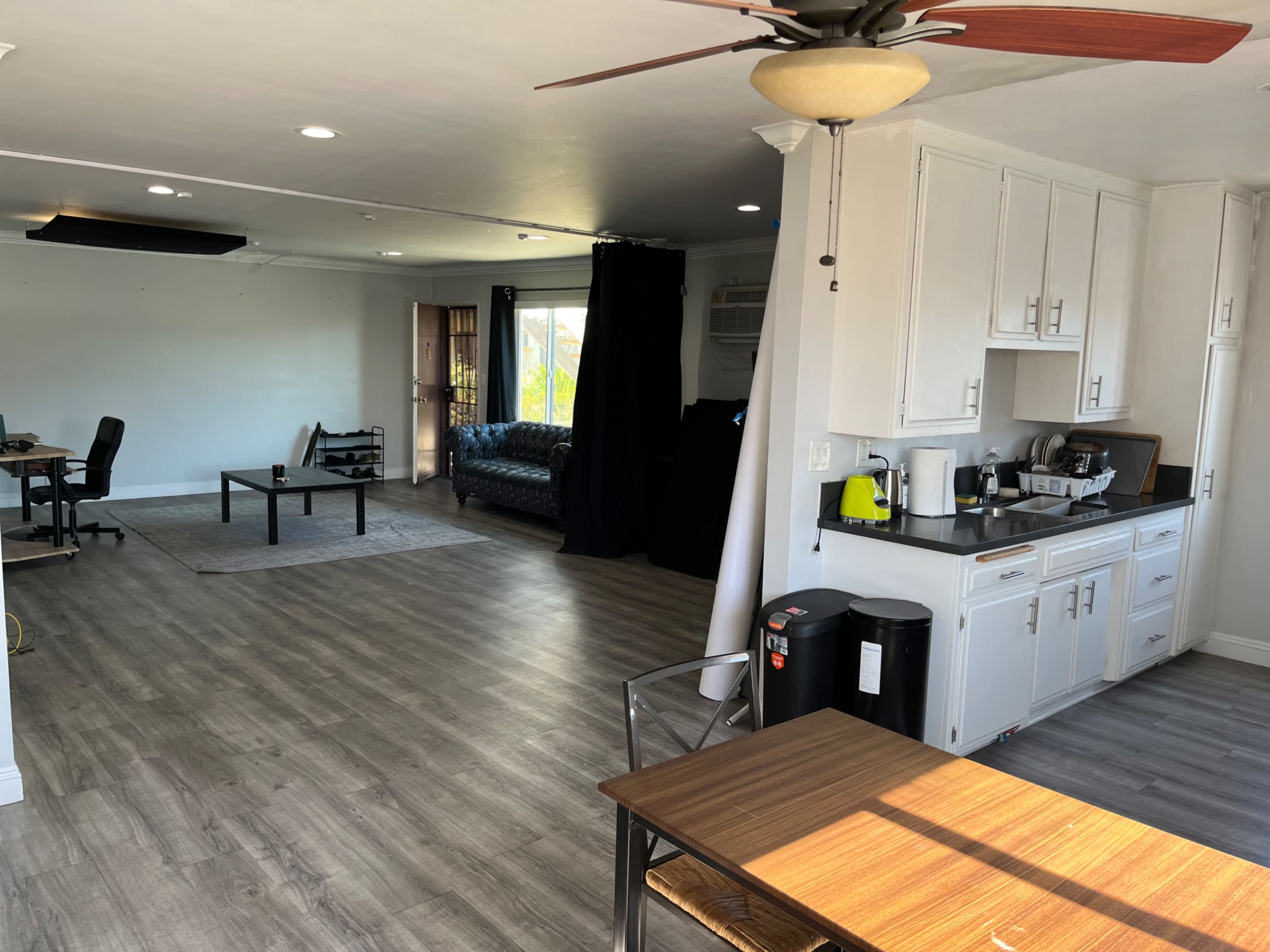 The image shows a spacious living area with a kitchen in the foreground, featuring a dining table, a coffee maker, and a couch in the background, along with workspaces on one side.