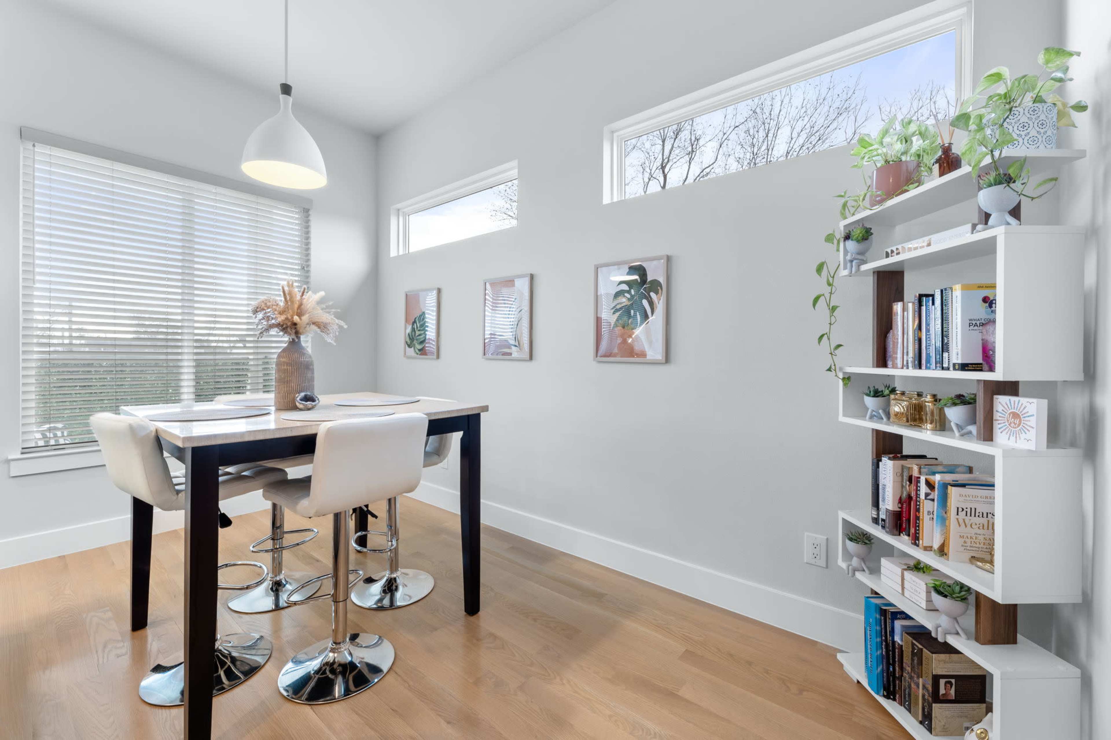 A modern dining area features a table with three stools, large windows allowing natural light, and a bookshelf lined with plants and books.
