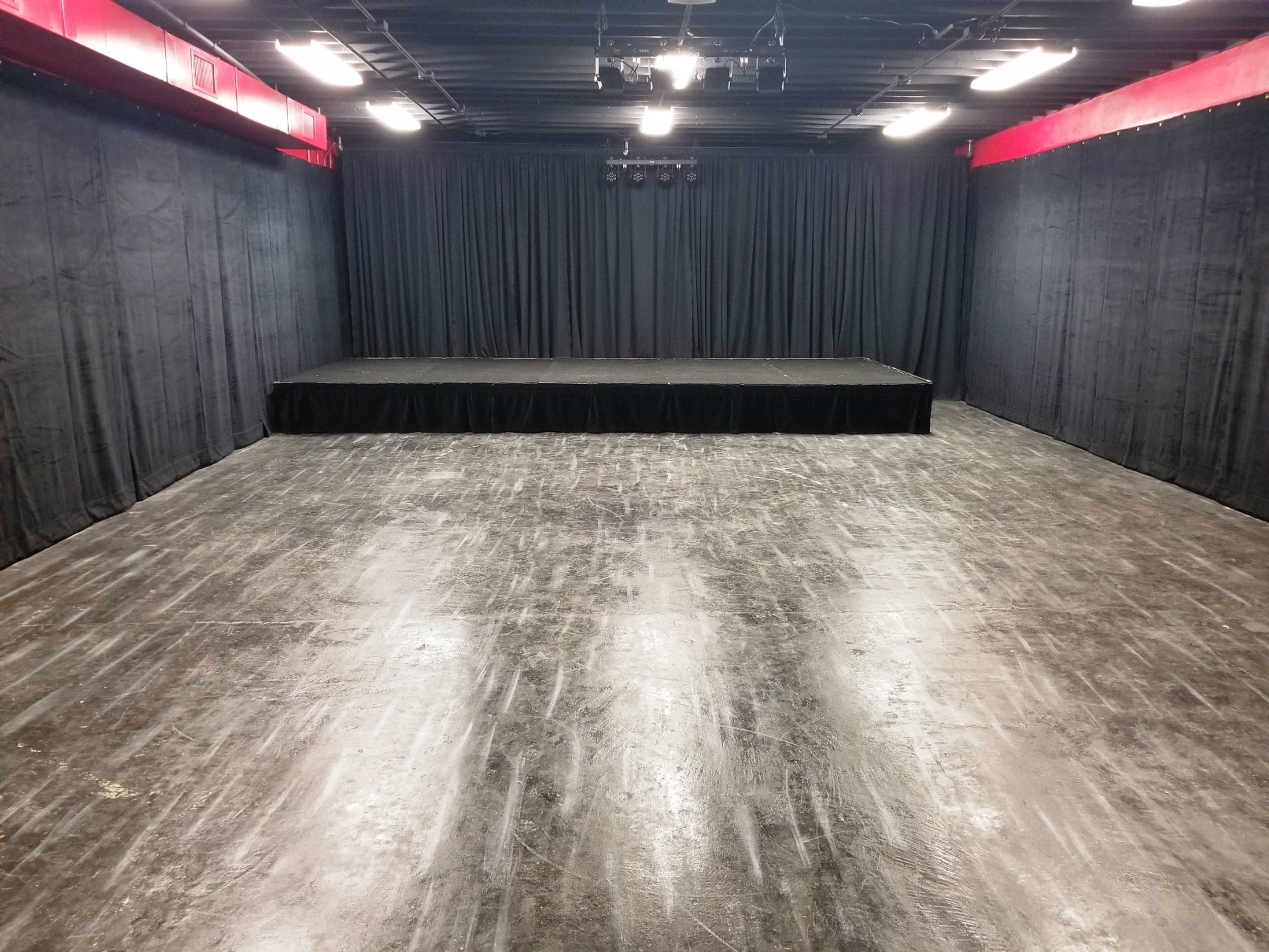 The image shows an empty performance space with a raised stage and black curtains in a dimly lit room.