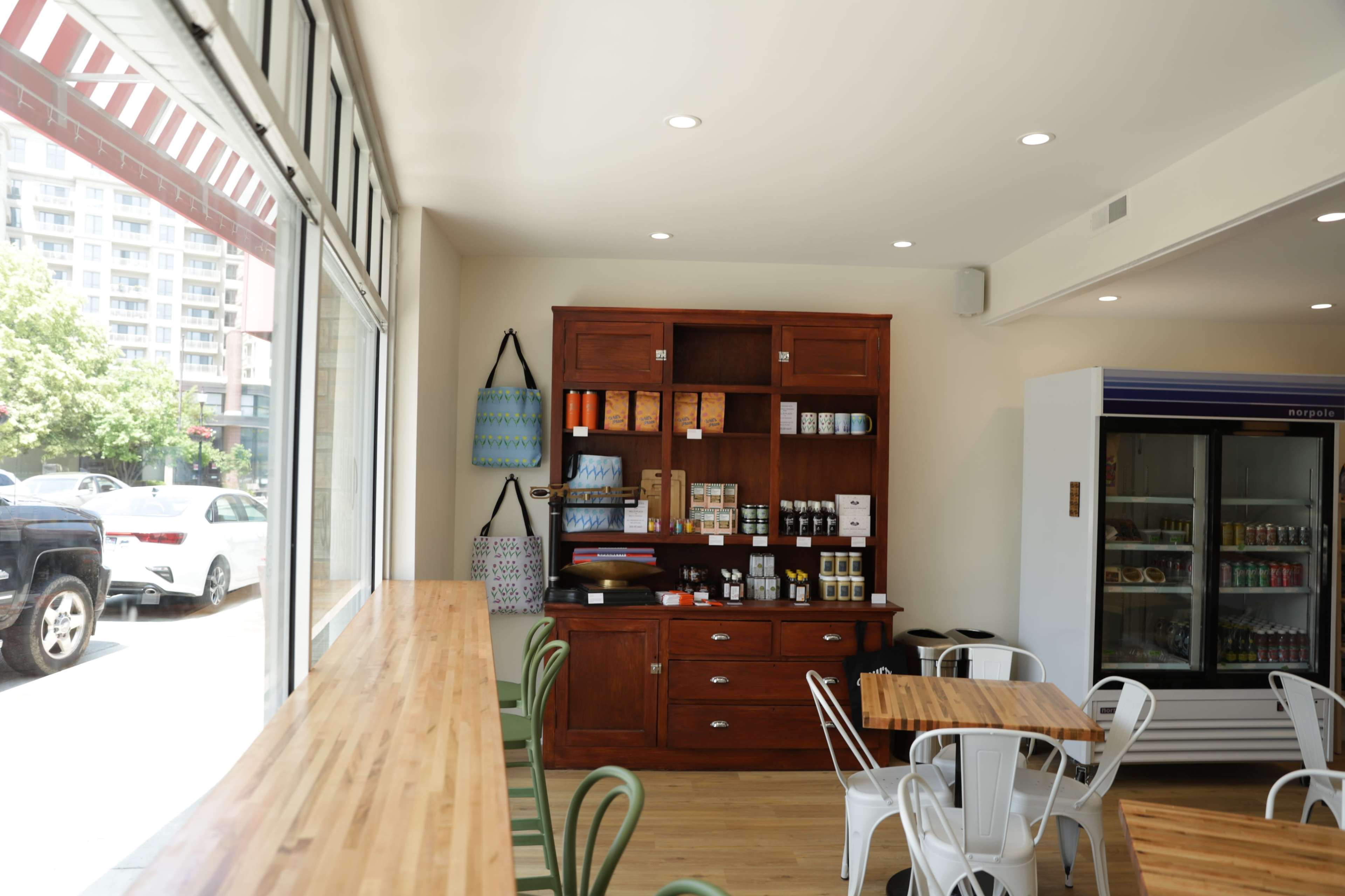 A modern café interior features a wooden countertop, a display shelf with various products, and metal seating arranged around tables.