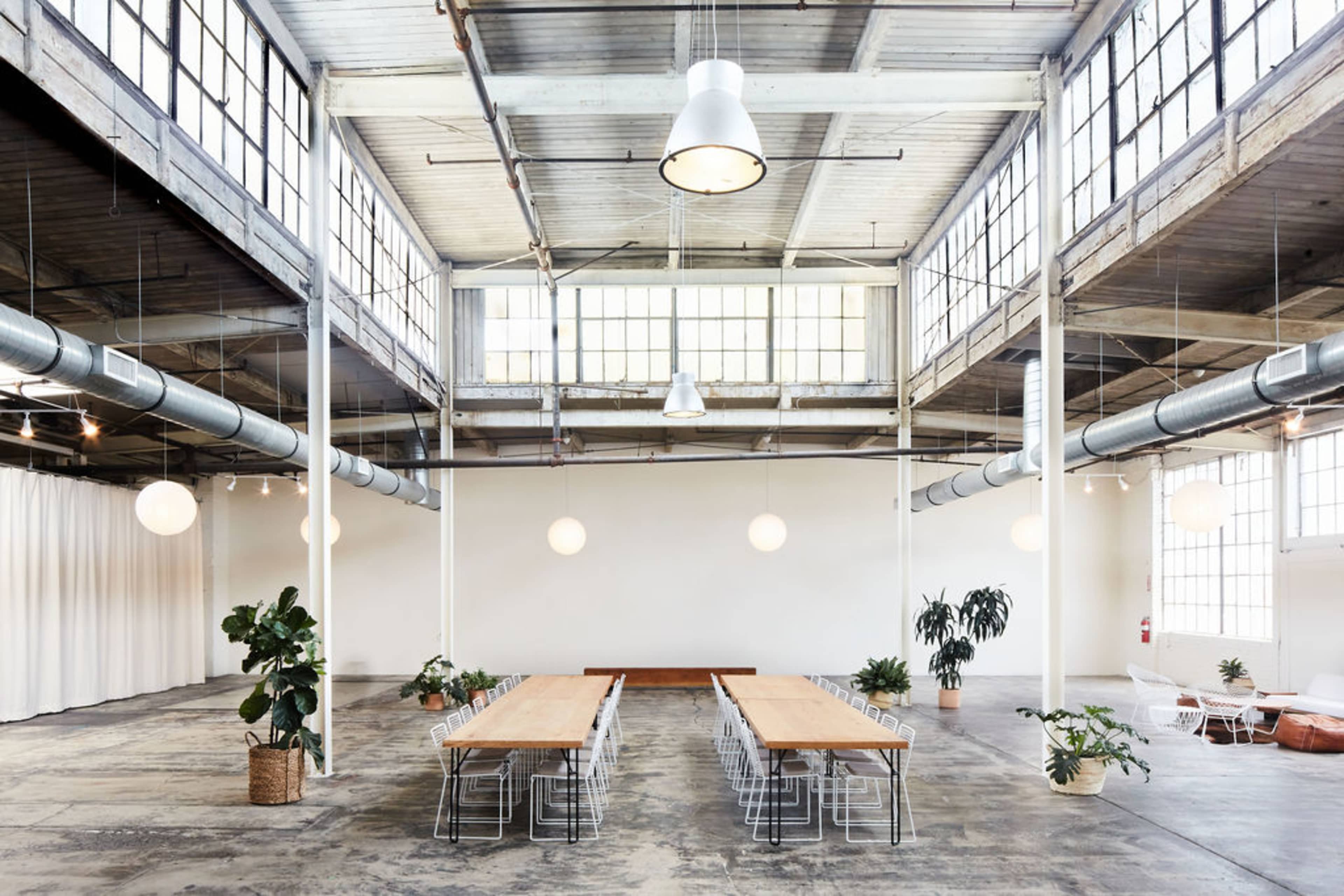 A large, open space features a long wooden table surrounded by metal chairs, with tall windows and plants positioned throughout the room.