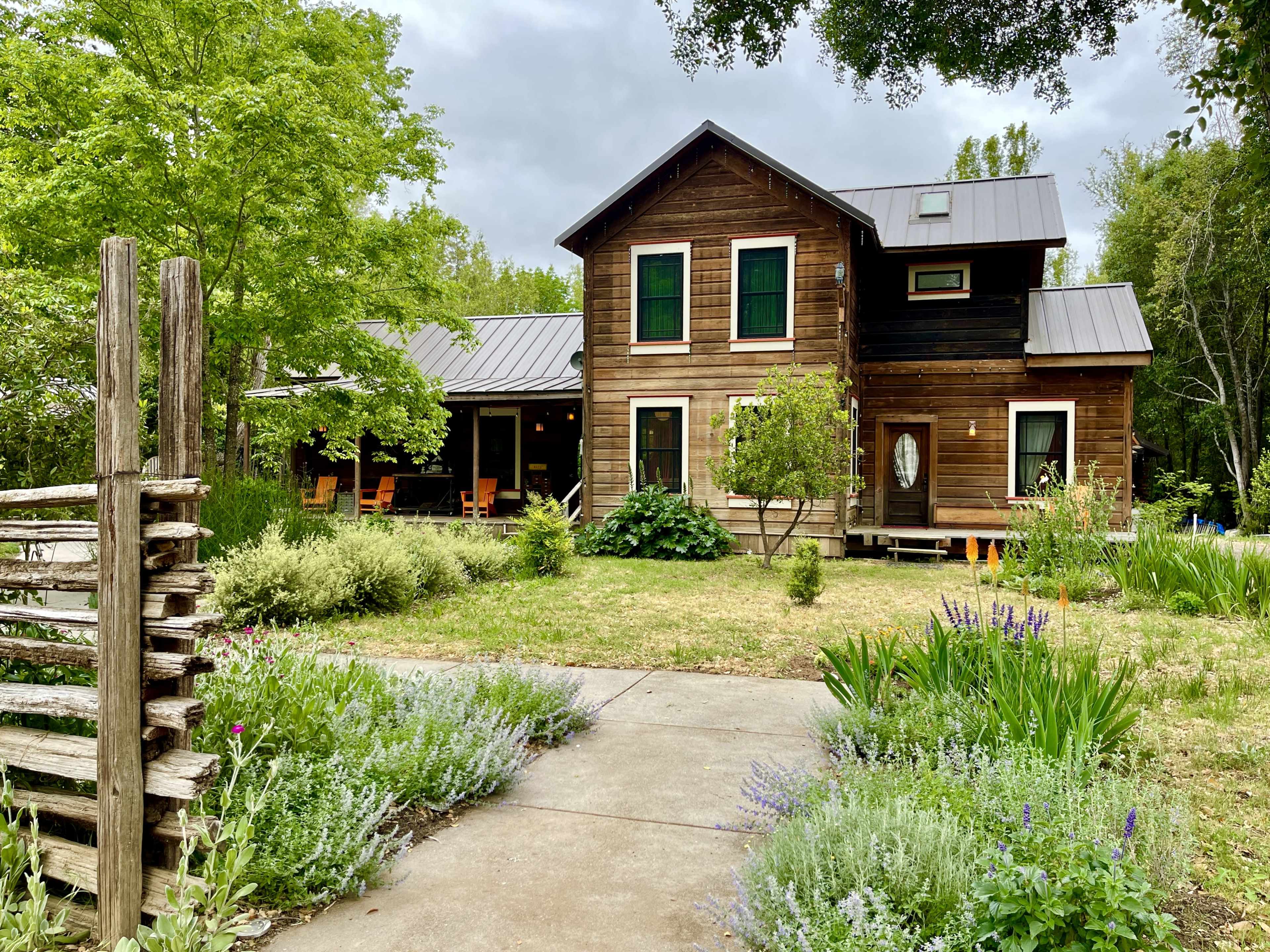 A rustic wooden house with a metal roof is surrounded by greenery and a gravel pathway.