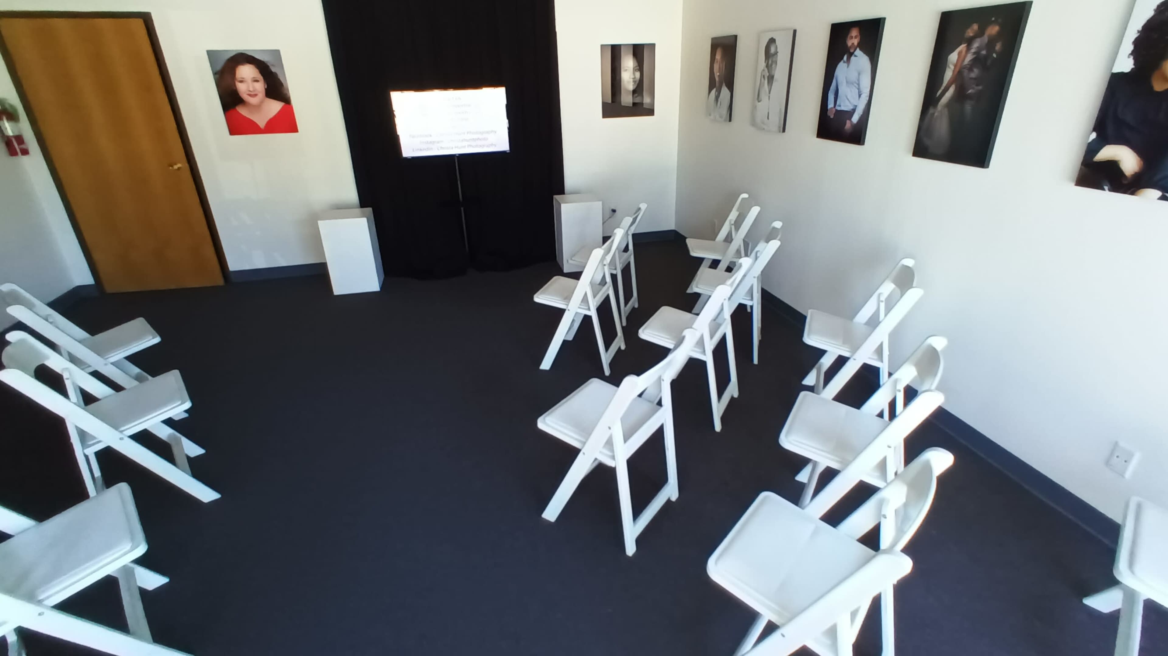 The image shows a room set up with white folding chairs arranged in rows facing a screen, with a black curtain in the background and framed portraits on the walls.