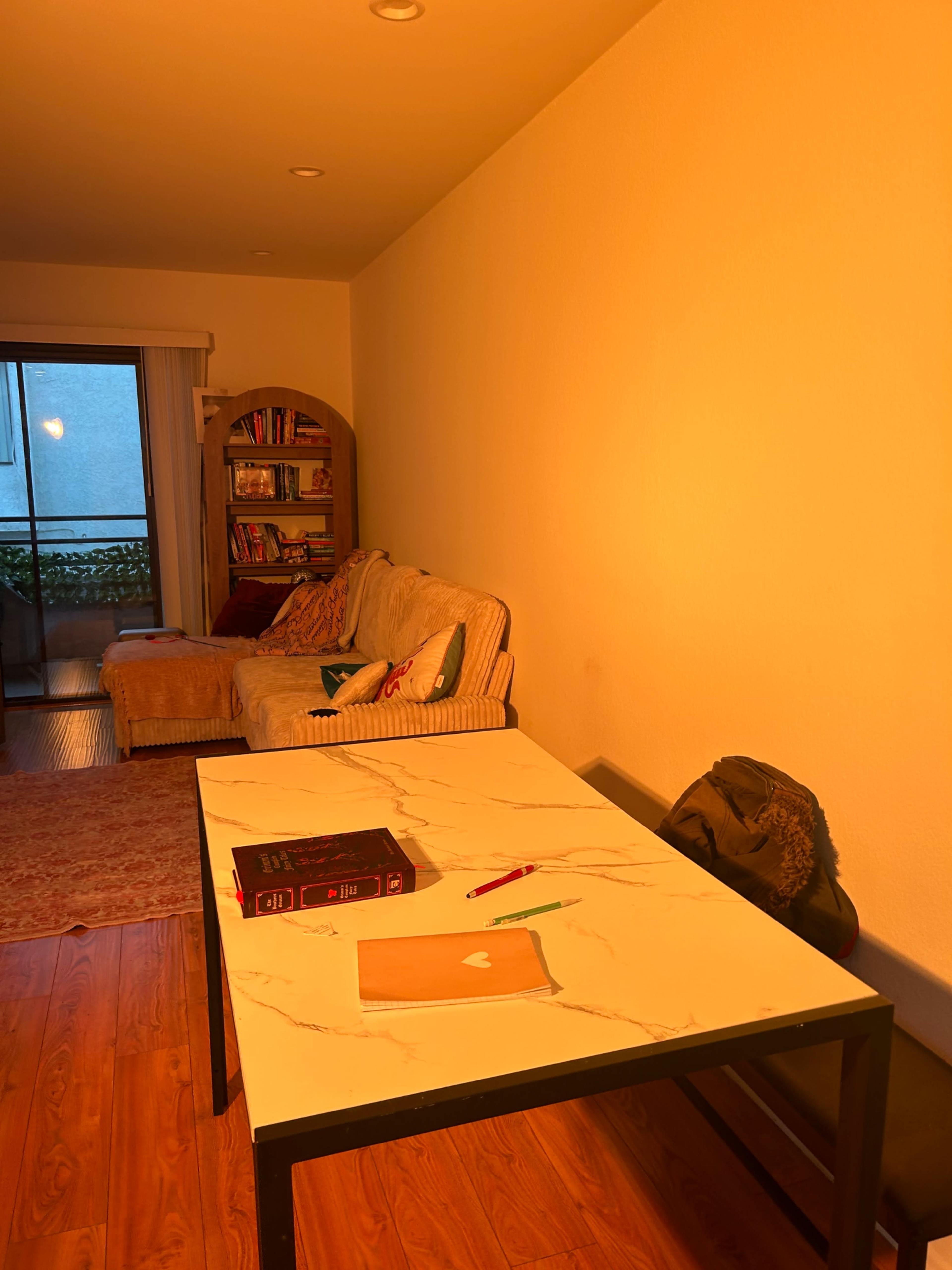 A small living space features a couch, a bookshelf, and a table with a notebook and pens, illuminated by warm light.