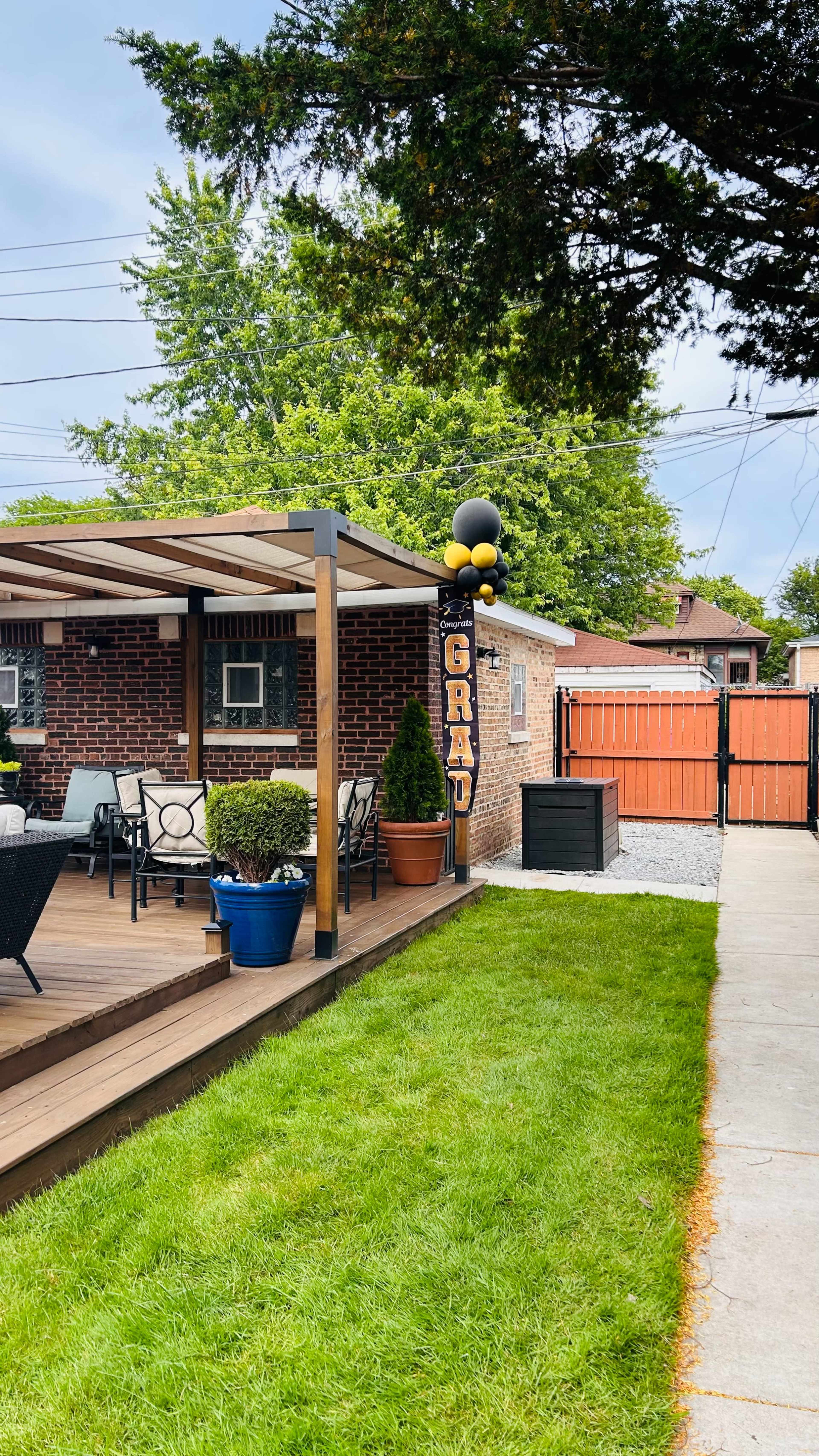 A backyard features a wooden deck with seating, adorned with a "GRAD" balloon display beside a pathway leading to a fenced area.