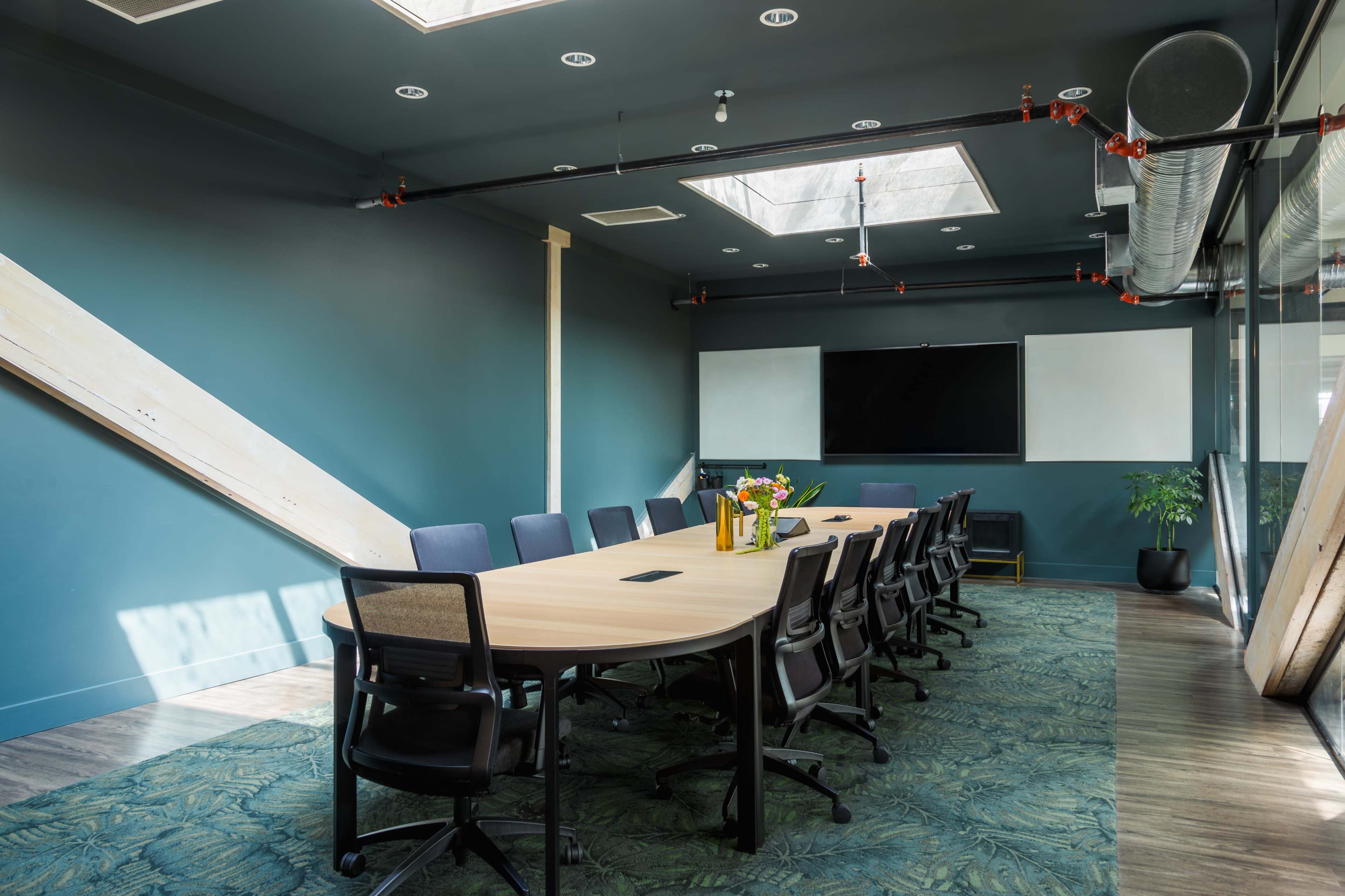A modern conference room features a long table surrounded by ergonomic chairs, with large windows and a flat-screen display on one wall.