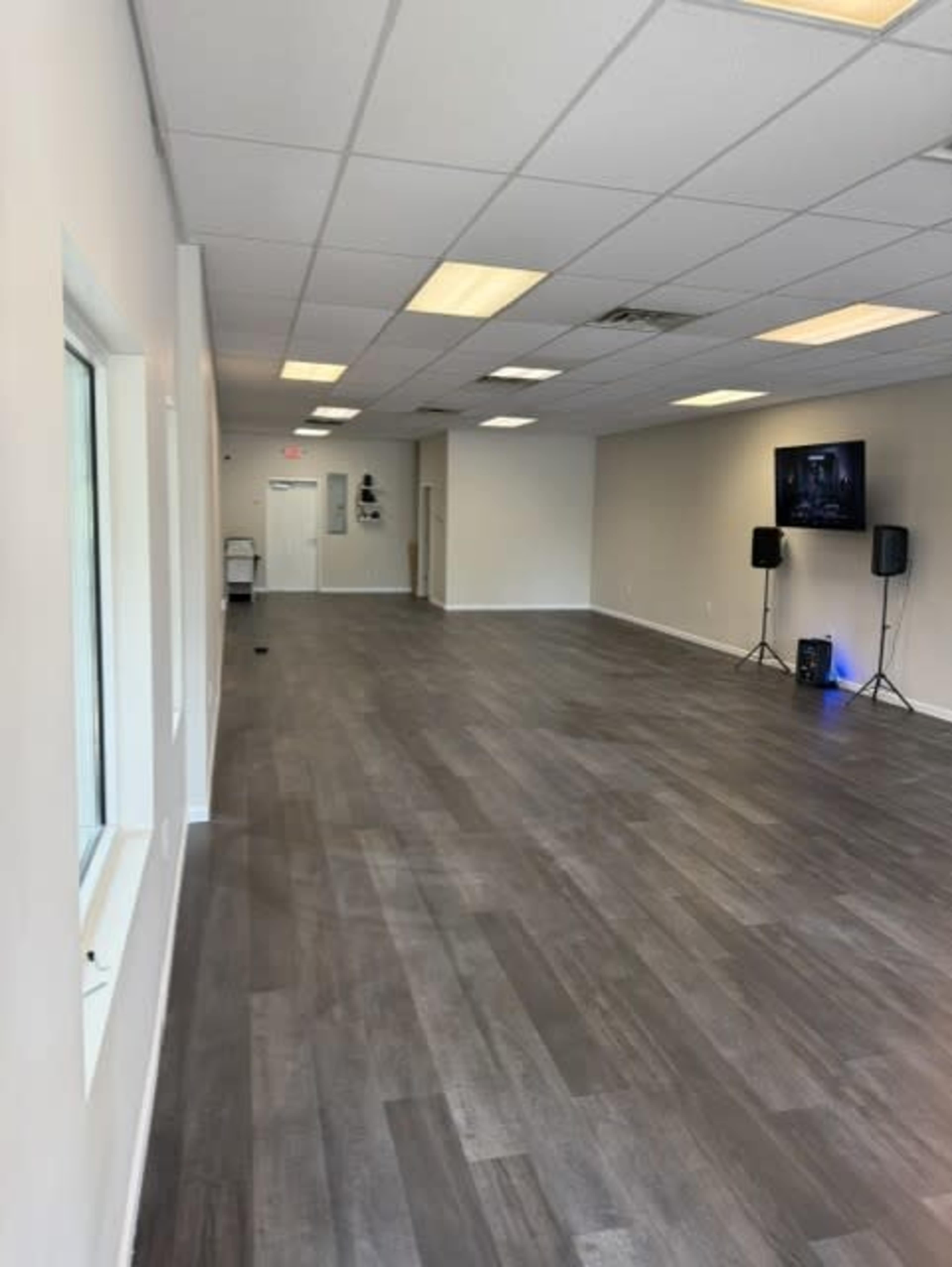 The image shows a spacious, empty room with light-colored walls and large windows, featuring a polished wooden floor and two speakers positioned along one side.
