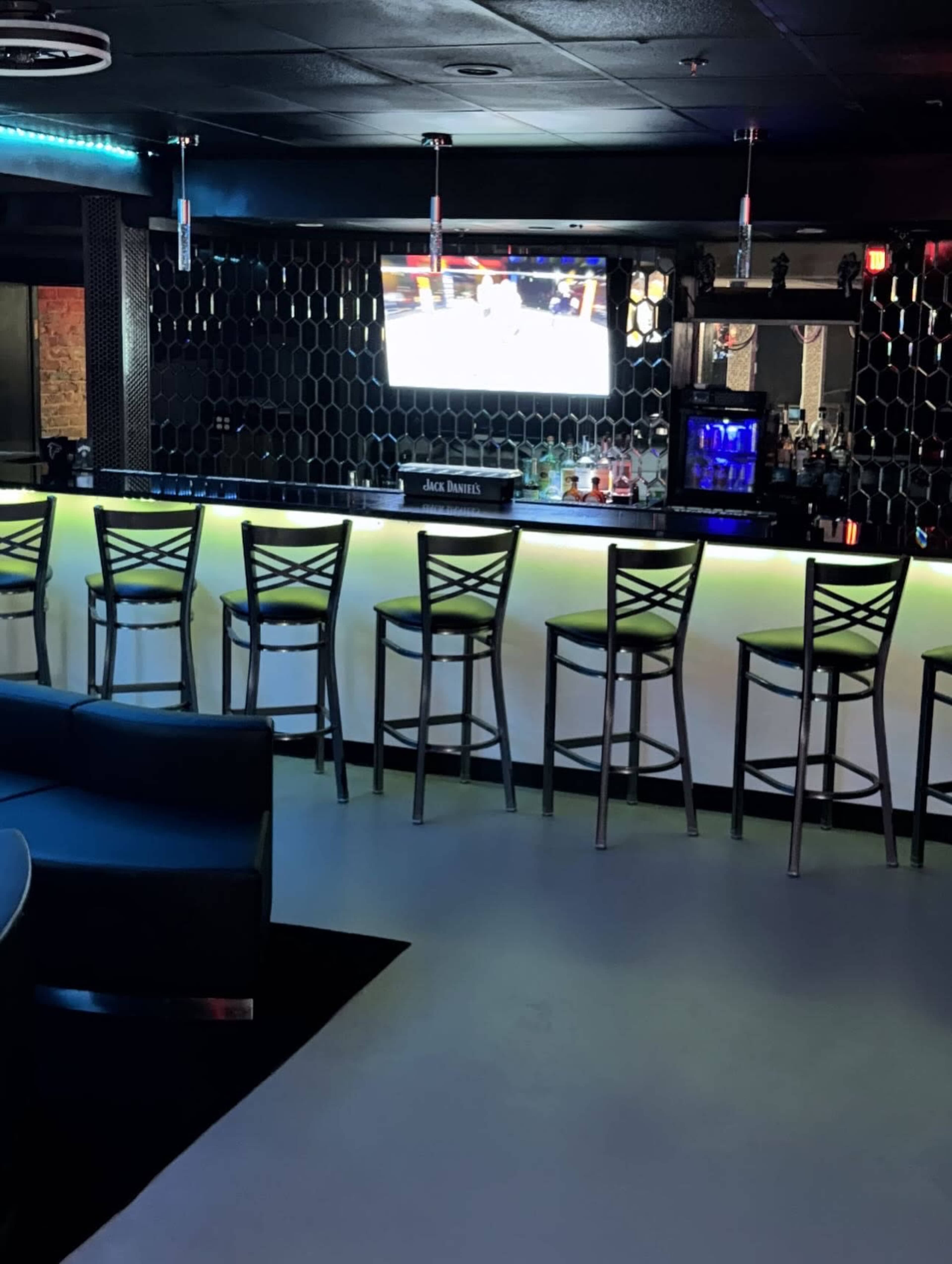 The image shows a dimly lit bar area featuring a long illuminated counter with several high chairs and a television mounted on the wall displaying a sports event.