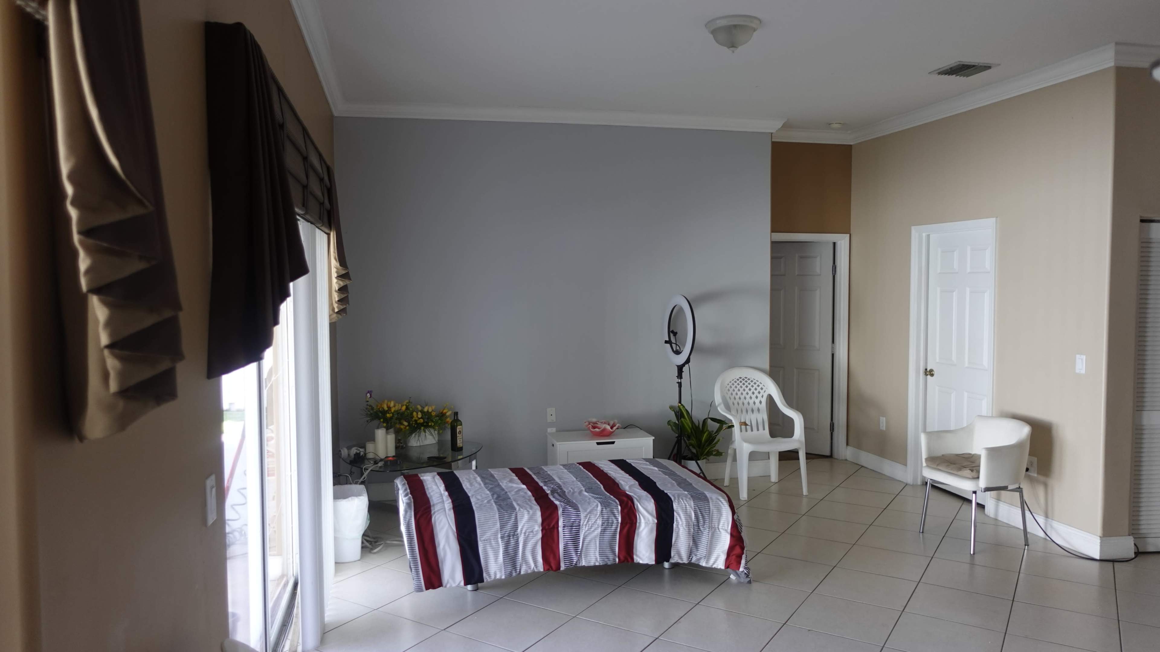 A spacious room features a bed covered with a striped blanket, a chair, and a ring light against a gray wall.