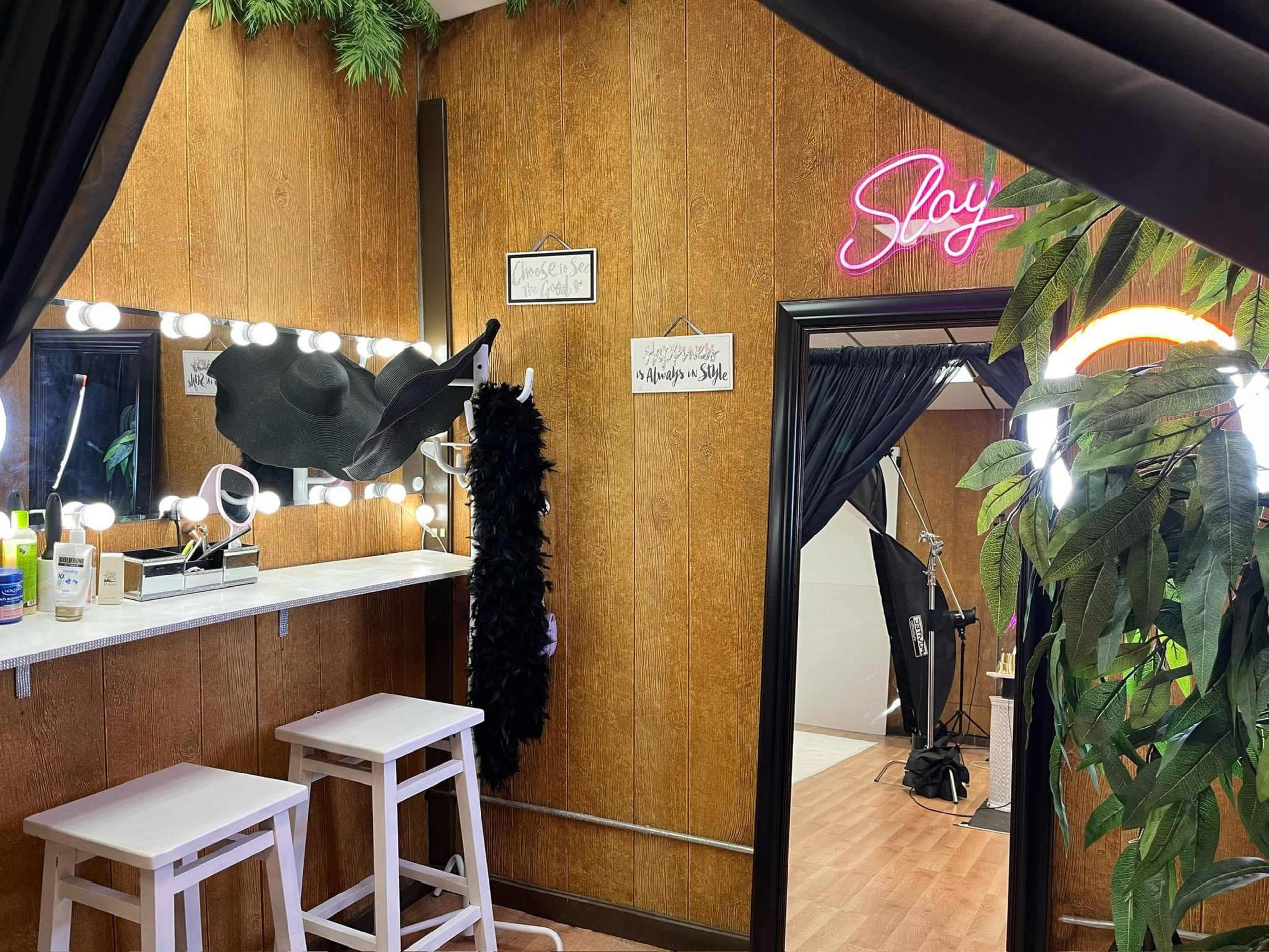 The image shows a makeup studio with a vanity mirror surrounded by lights, a large black feather boa hanging on a rack, and a neon sign that reads "Slay" on the wall.