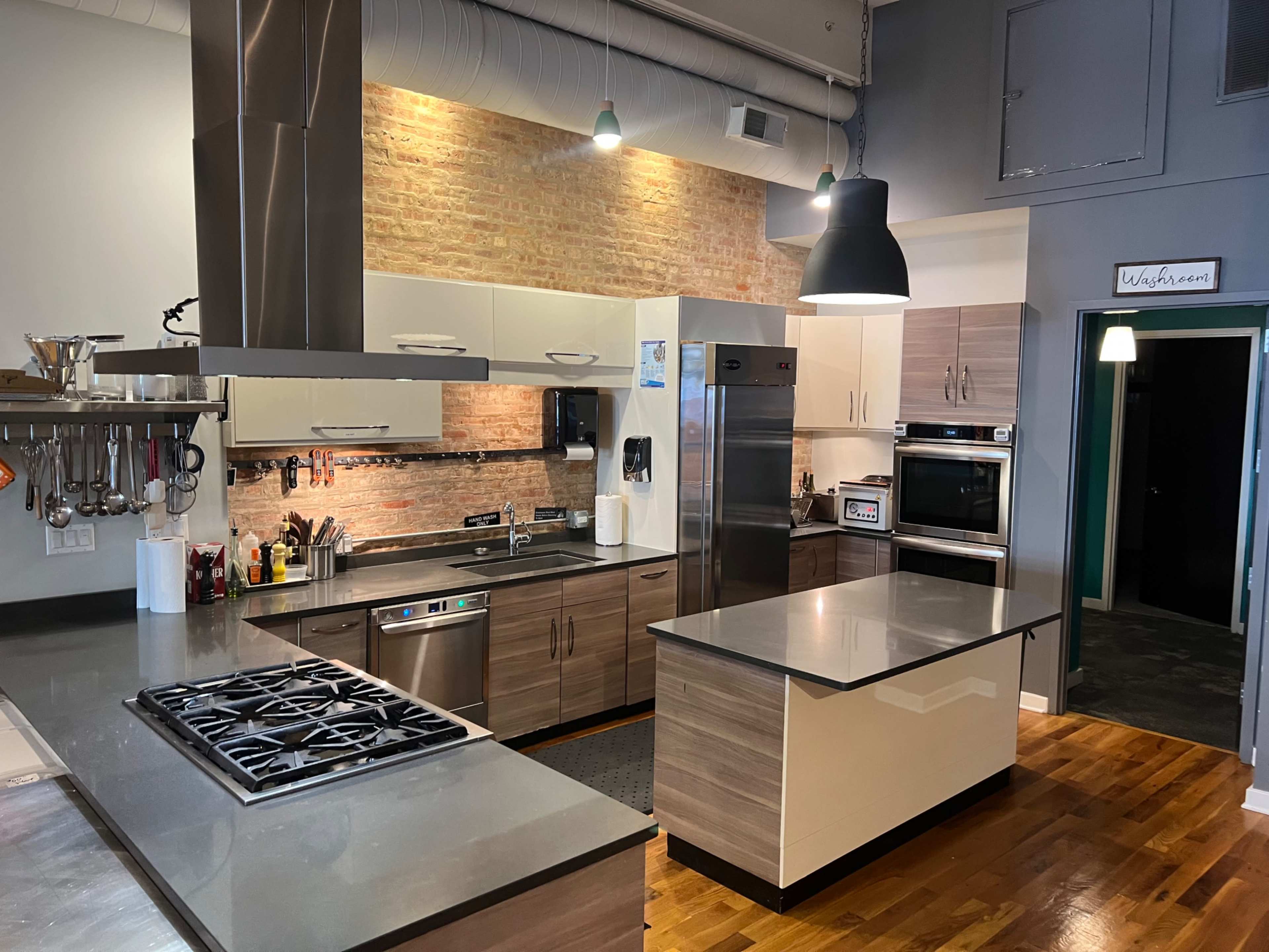 The image shows a modern kitchen with a central island, stainless steel appliances, and a brick wall backdrop.