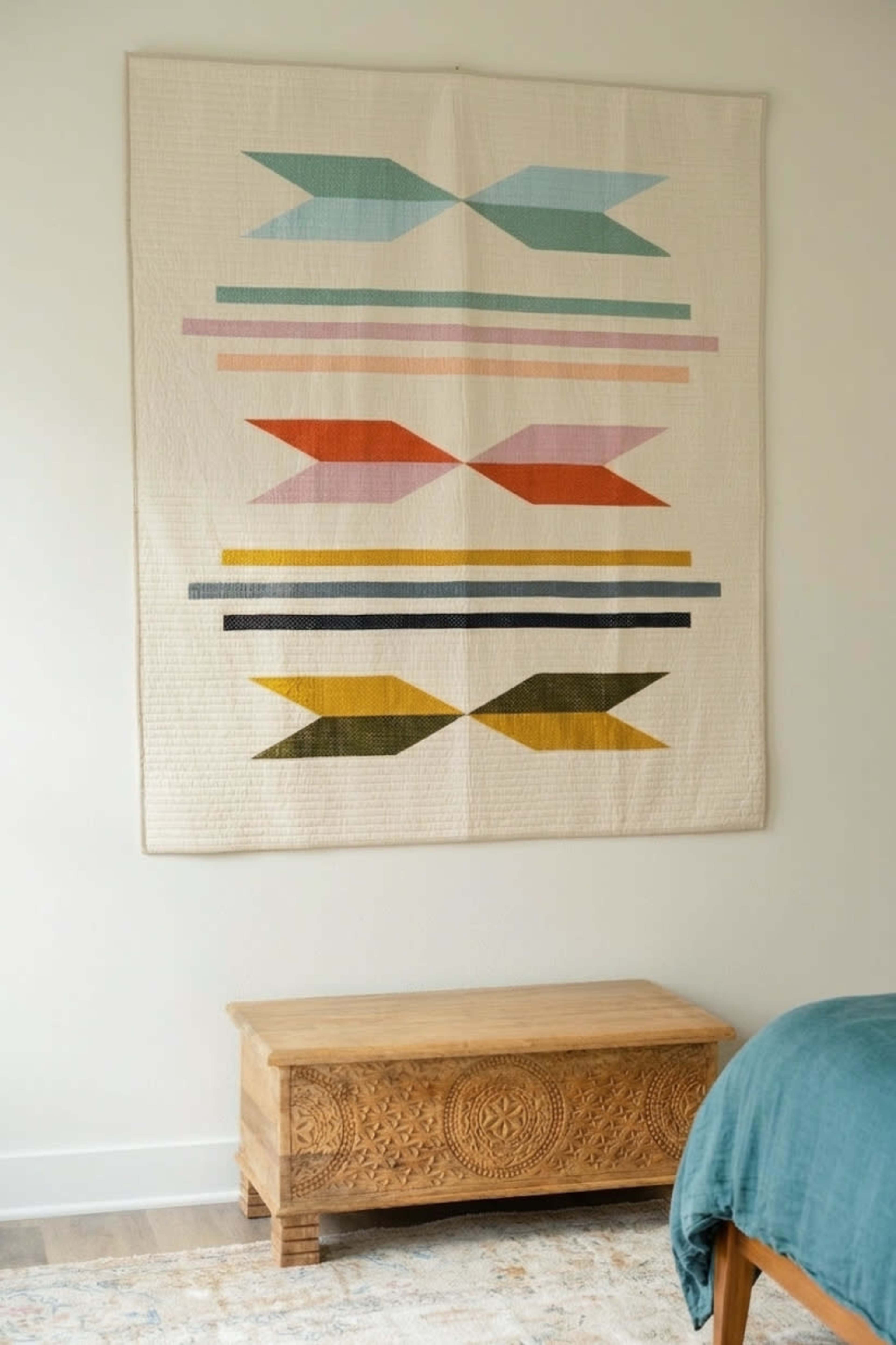 A geometric textile hangs on a wall above an intricately carved wooden trunk in a softly decorated bedroom.
