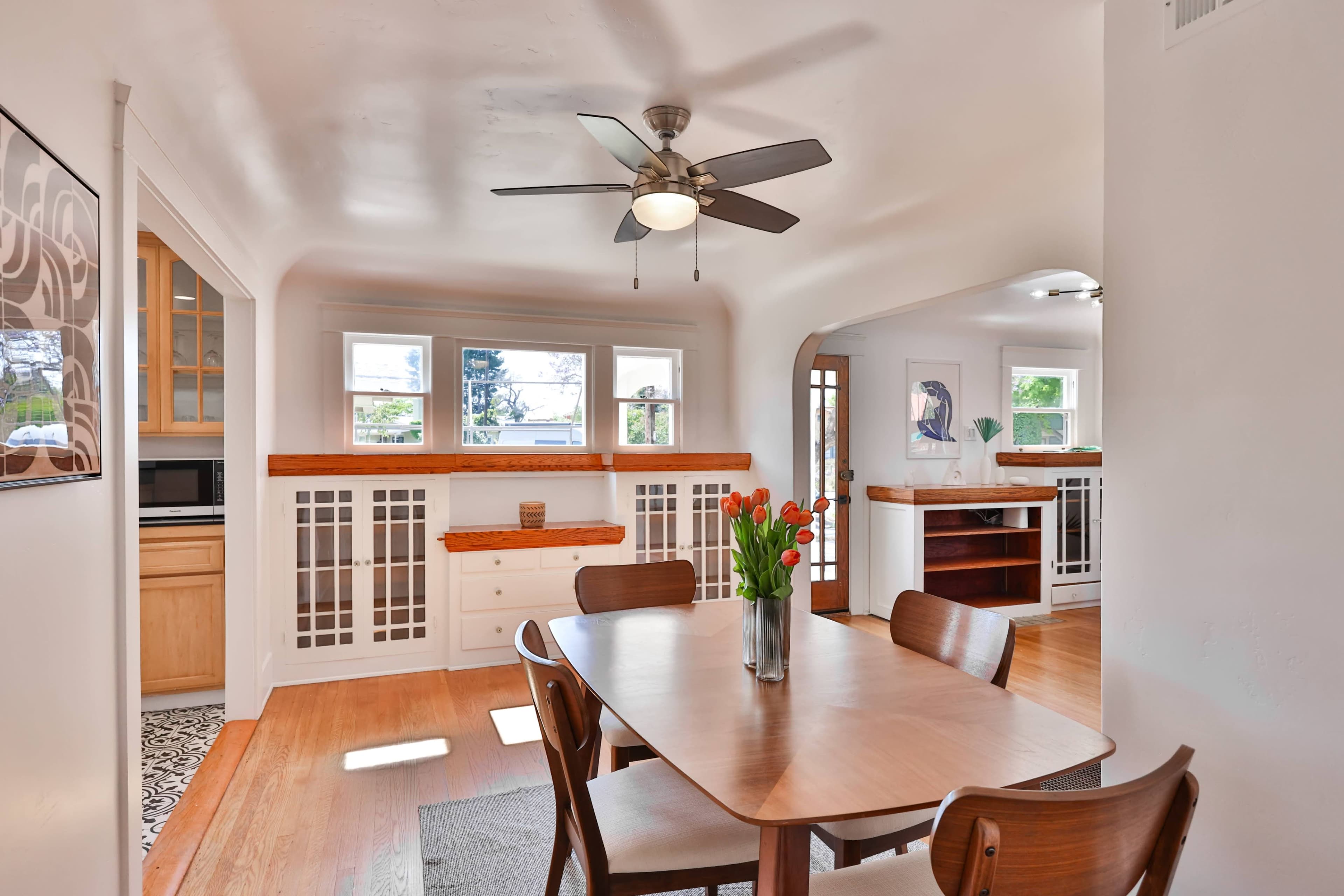 A dining area with a wooden table and chairs, featuring a ceiling fan and large windows, adjacent to a kitchen space.