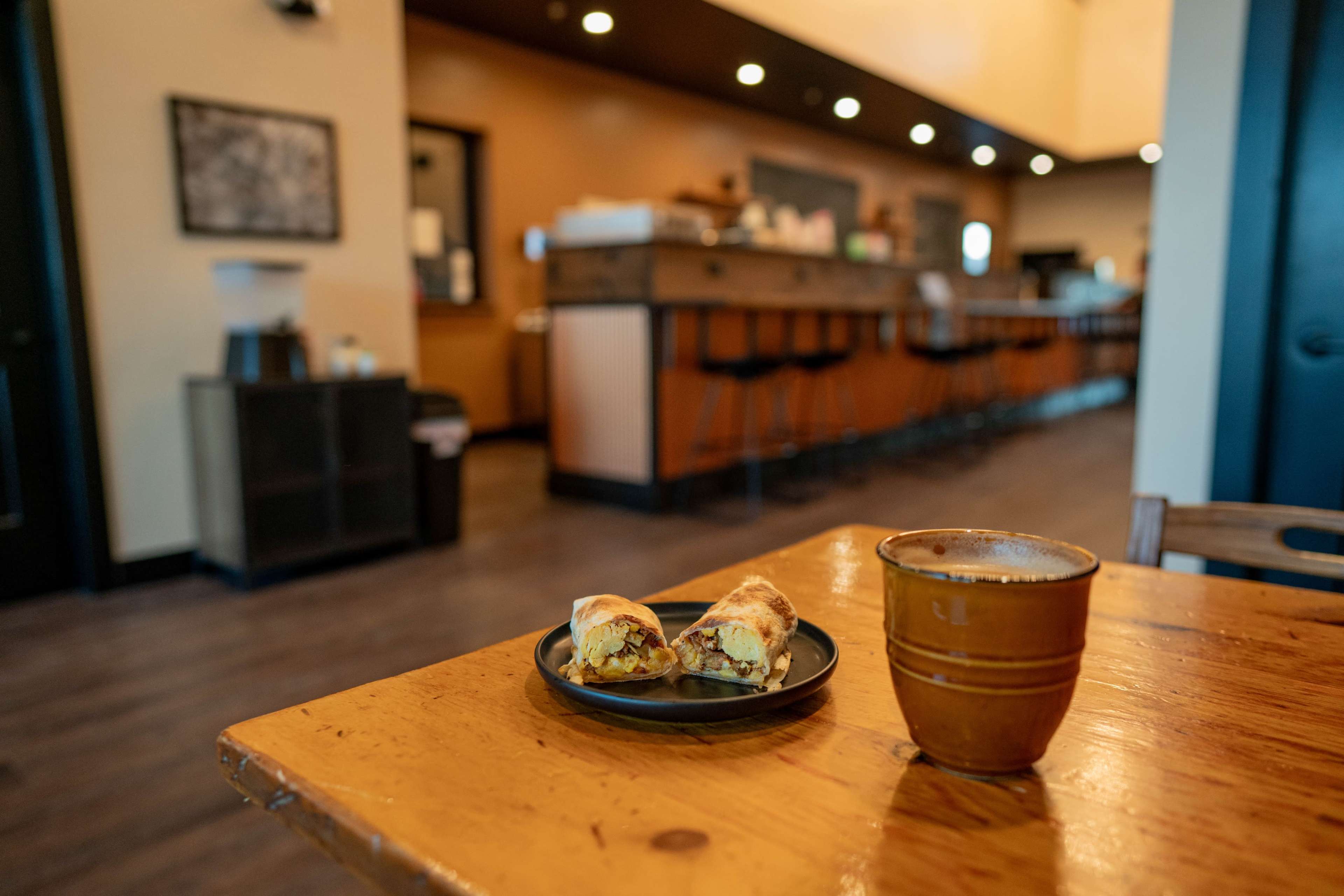 A plate with a burrito and a cup is placed on a wooden table in a casual café, with a bar area visible in the background.