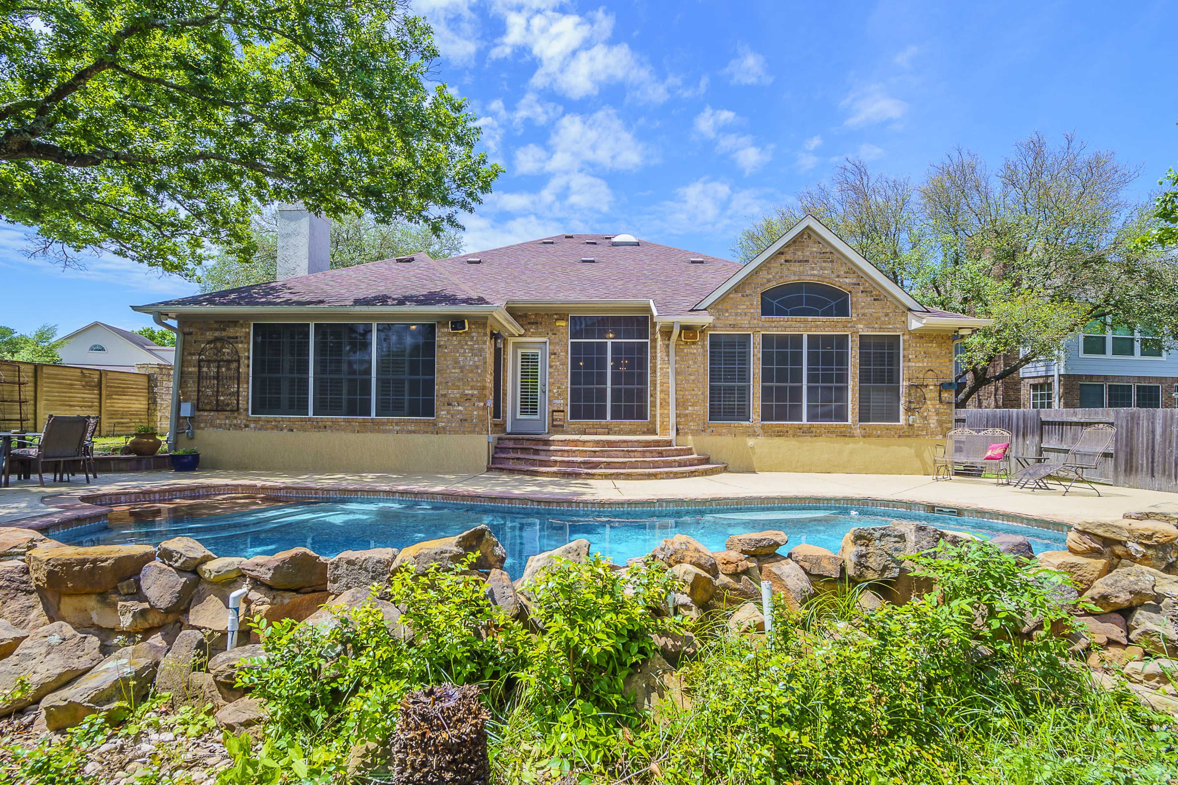 A single-story stone house with large windows overlooks a backyard pool surrounded by a stone wall and lush greenery.