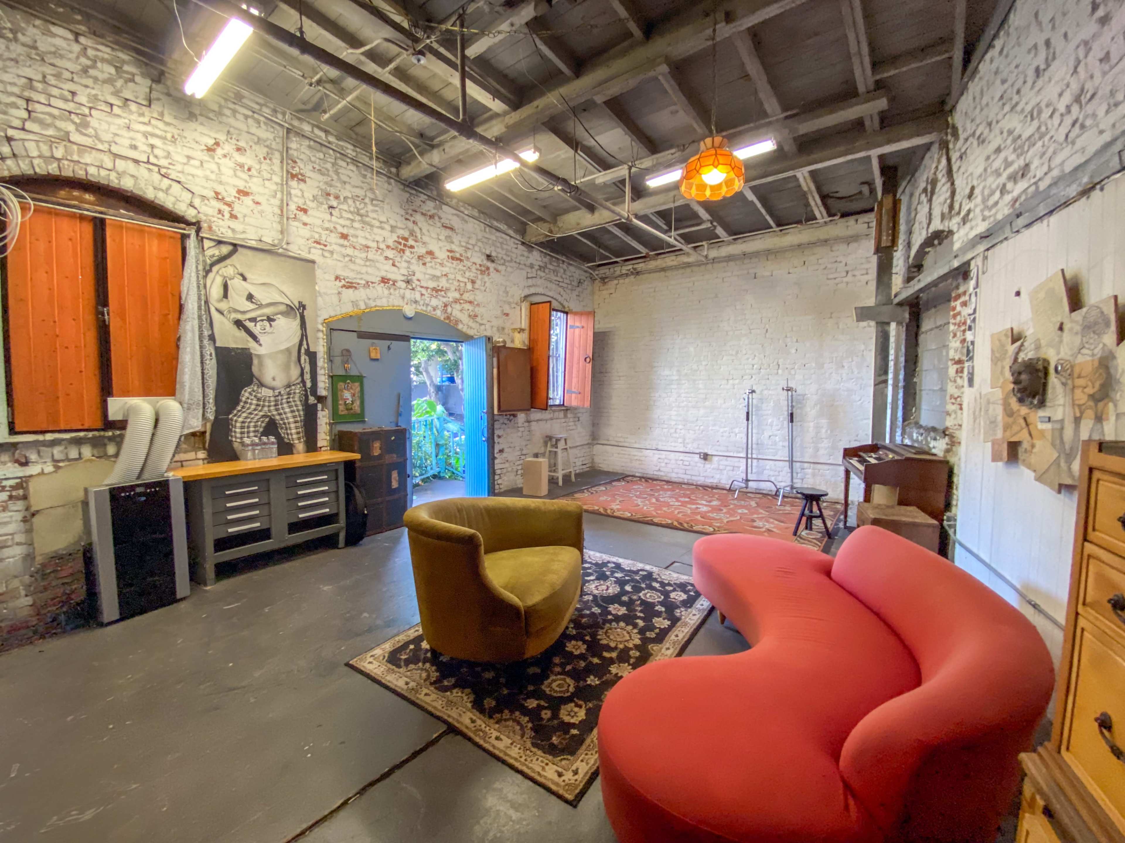 A spacious room with exposed brick walls, featuring a green and red modern couch, a patterned rug, and various furniture pieces scattered throughout.