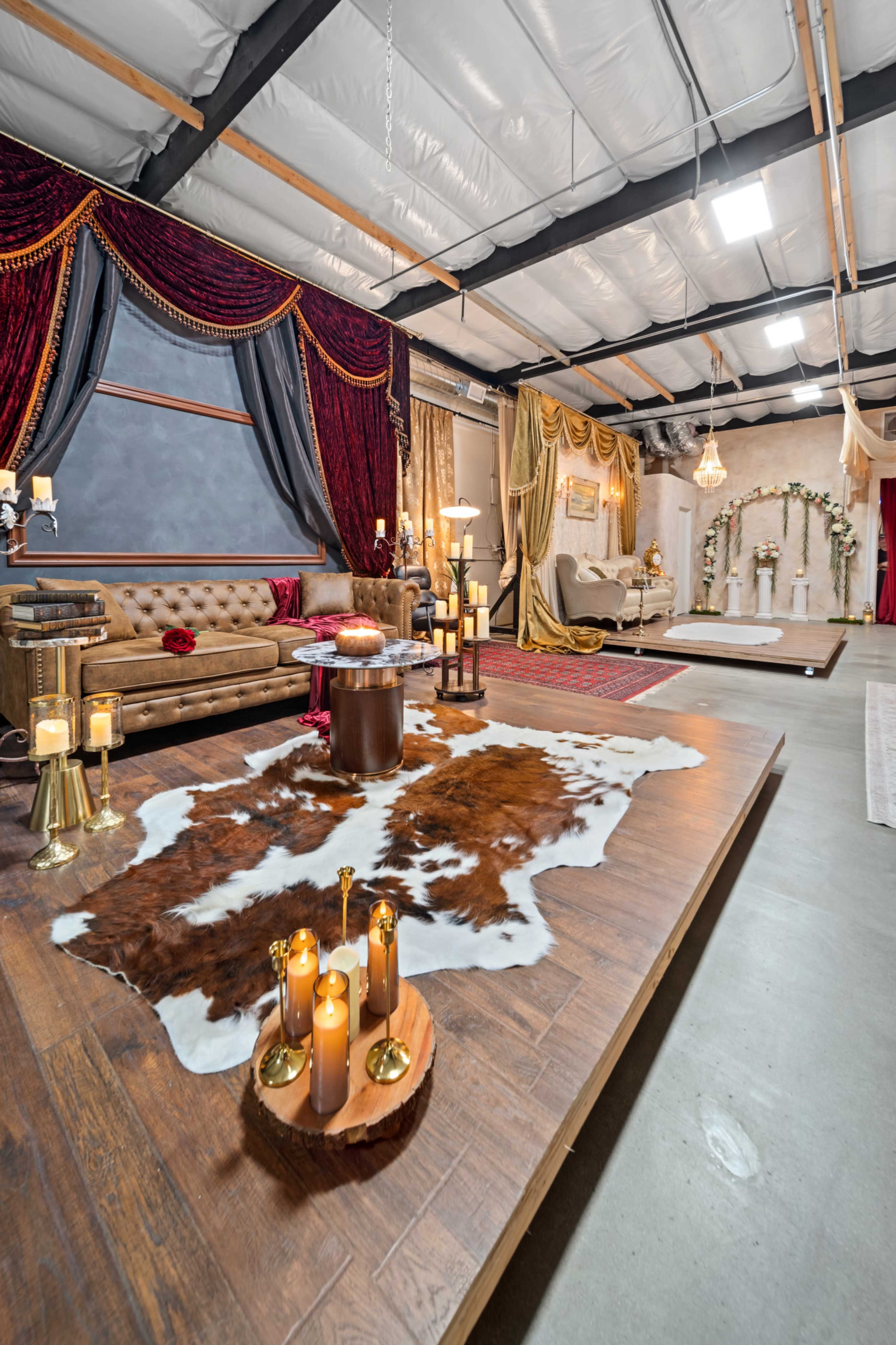 The image shows a spacious indoor space with elegant decor, including a large cowhide rug, candles on a wooden stand, a plush sofa, and various draped fabric backgrounds.