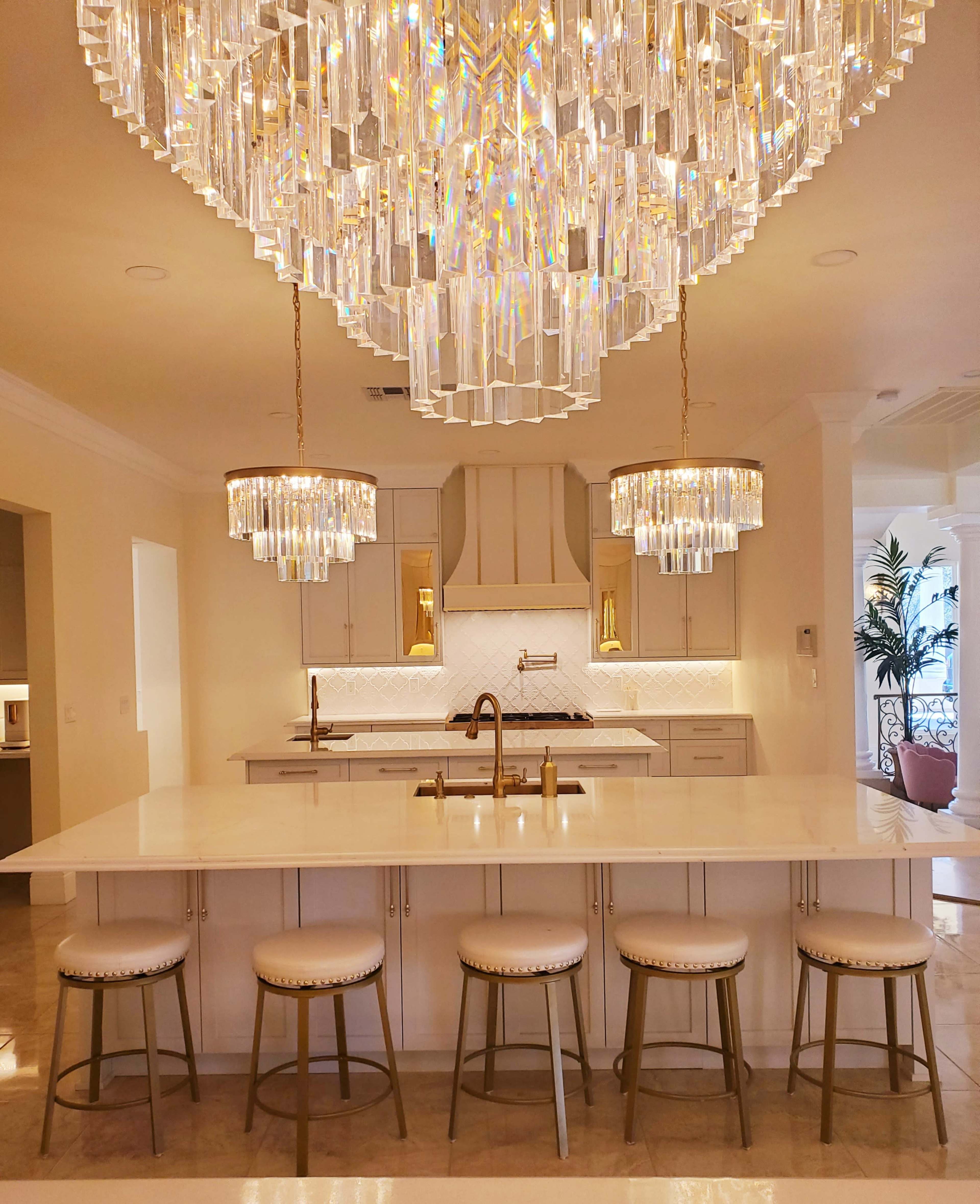 The image shows a modern kitchen with a large island, featuring gold accents, bar stools, and a sparkling crystal chandelier overhead.