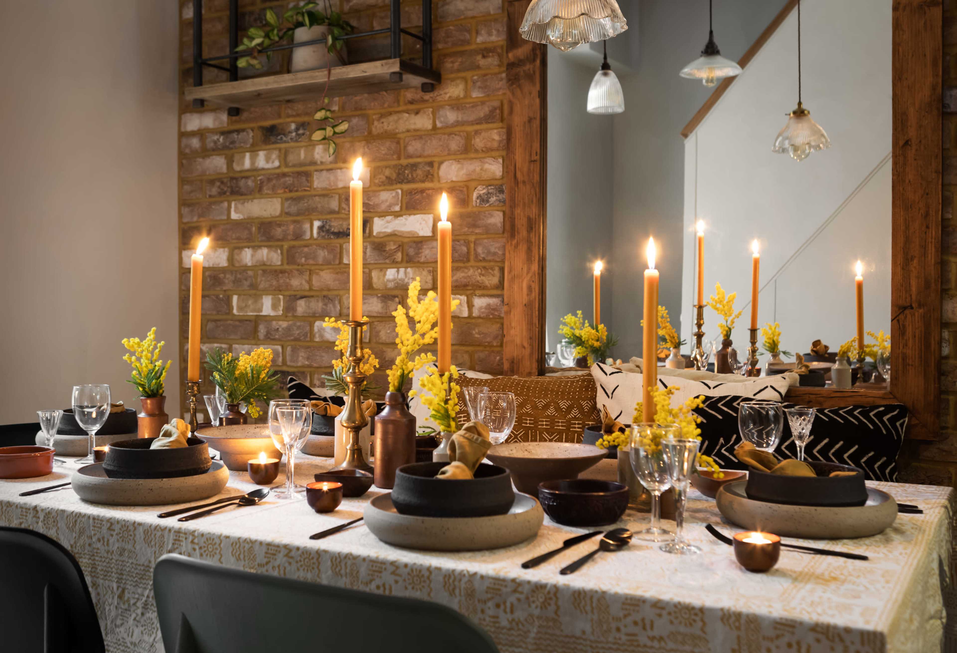 A dining table is set with candles, flowers, and various tableware against a backdrop of exposed brick and decorative lighting.