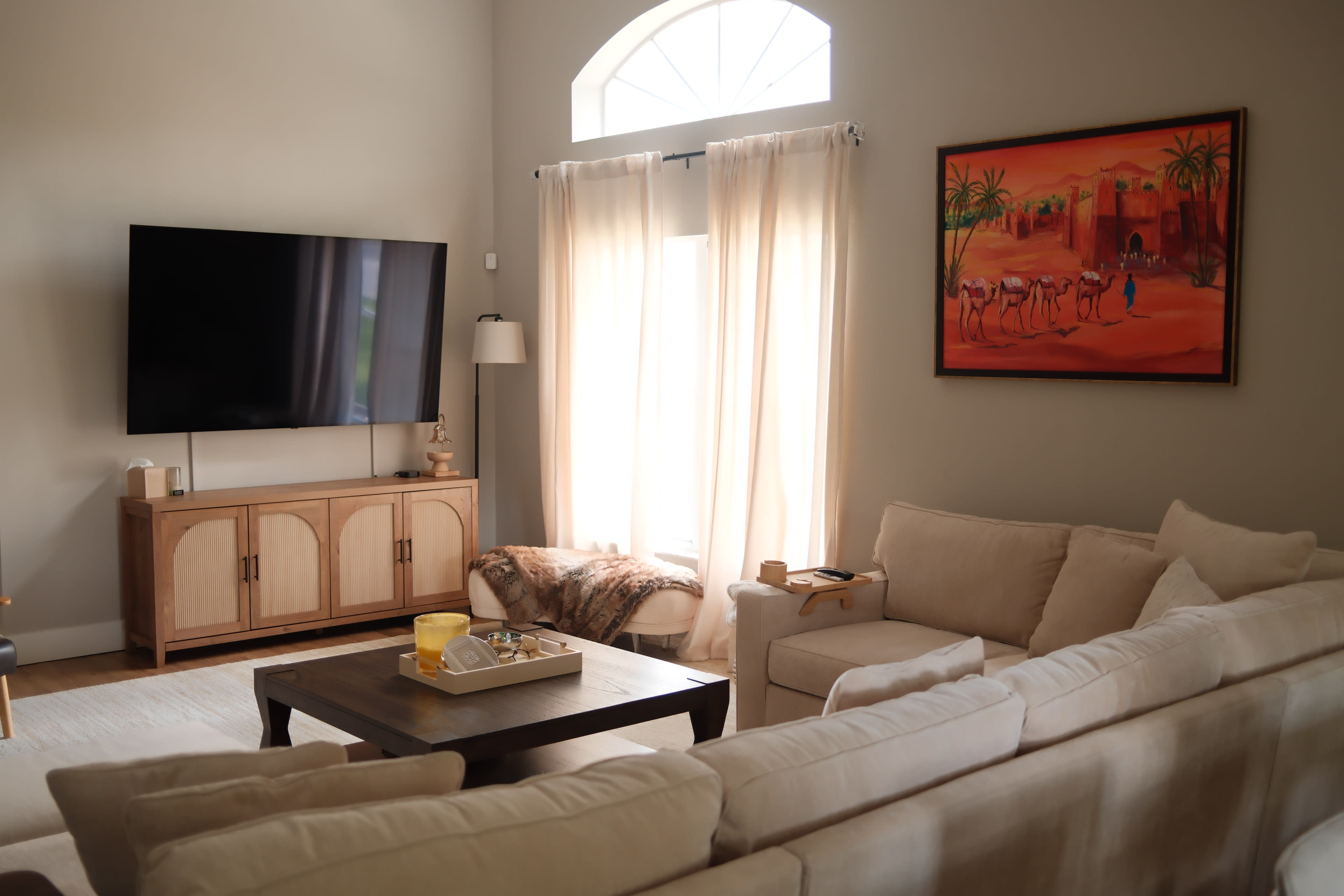 A spacious living room features a large TV mounted on the wall, a wooden entertainment center, a light-colored sofa, and a painting of a desert scene.