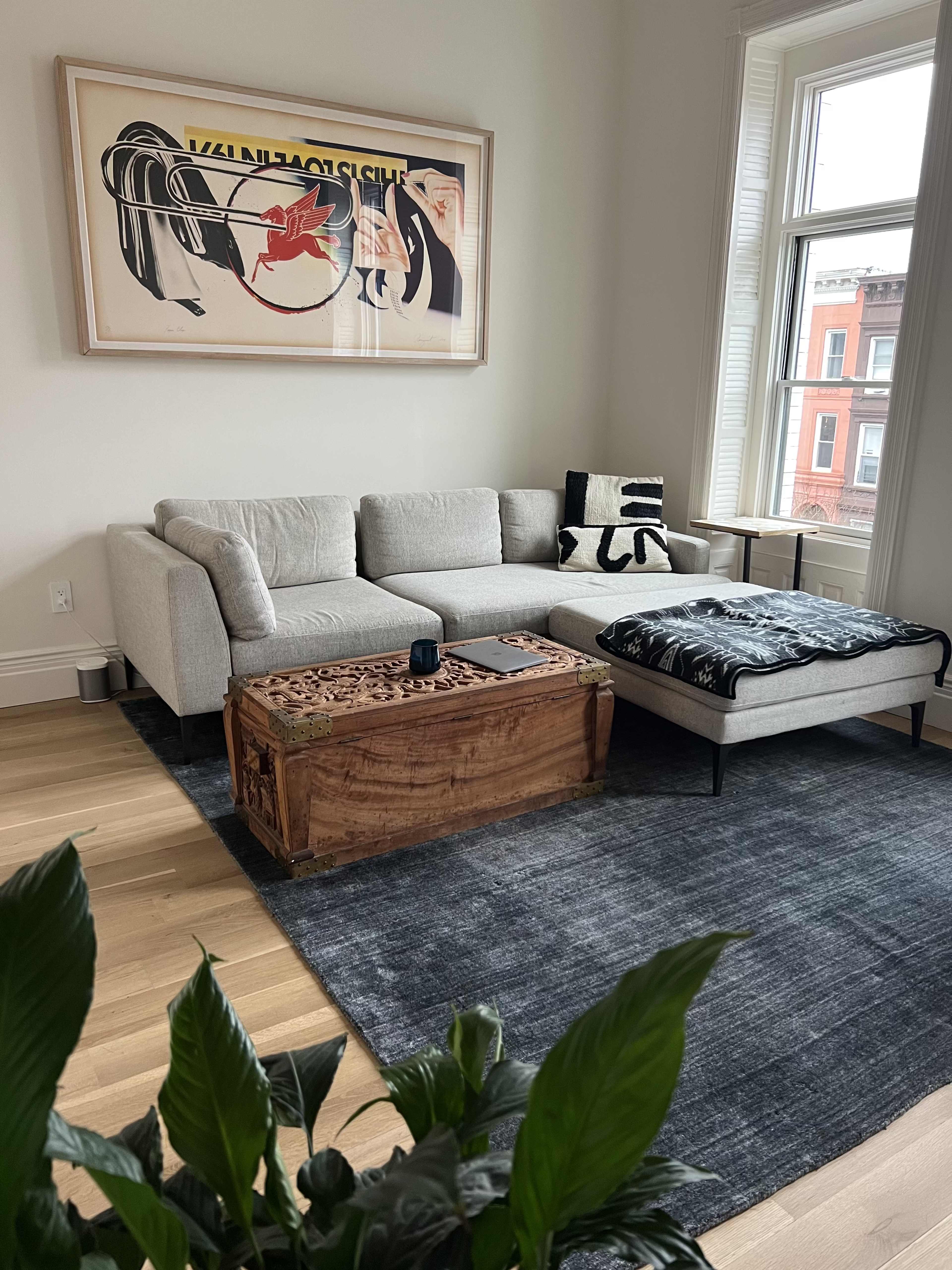 A light gray sectional sofa is positioned beside a wooden coffee table, with a black and white throw on a dark gray rug in a bright living room featuring a large artwork on the wall.
