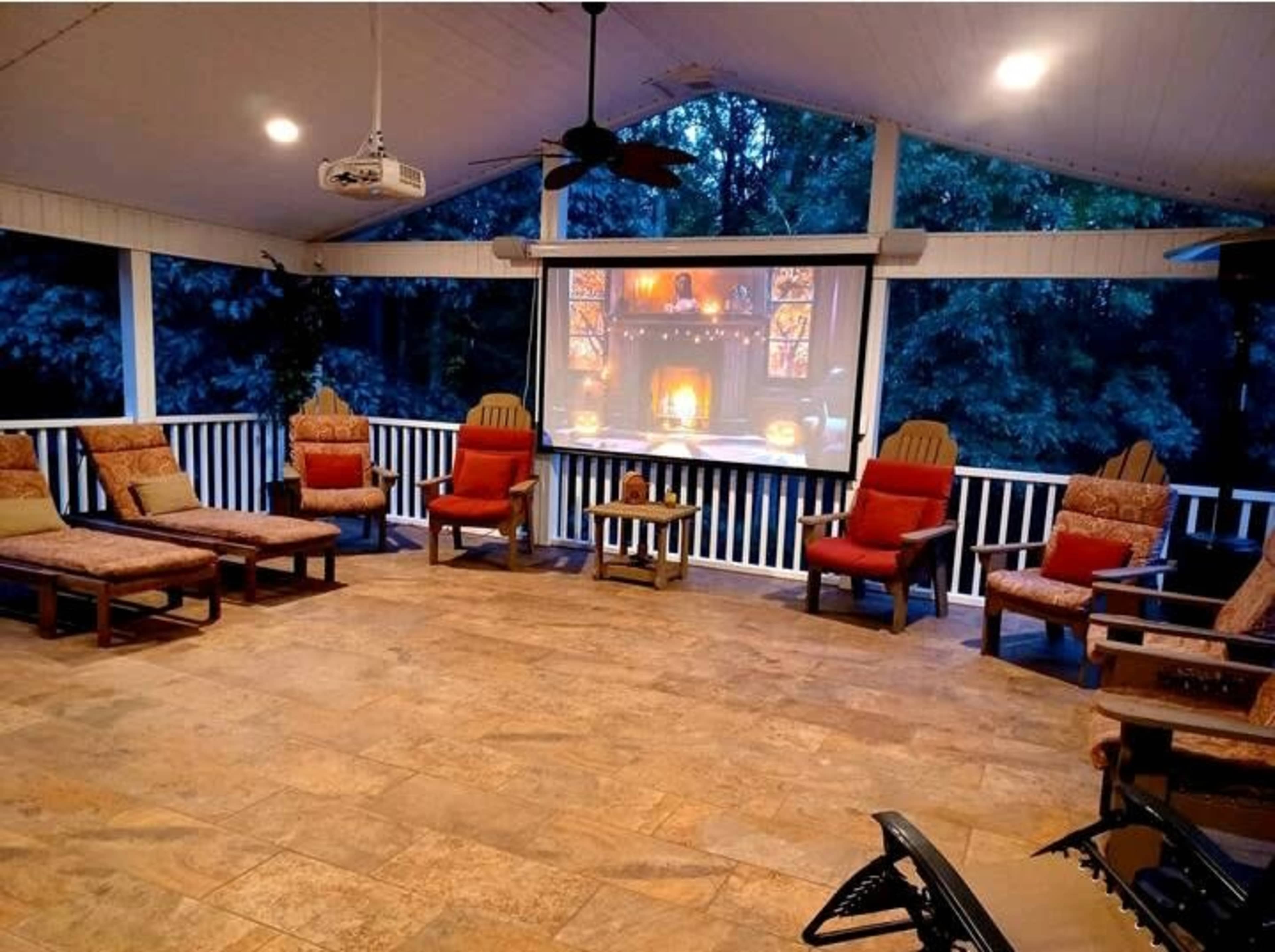 A spacious porch area with several wooden chairs and loungers arranged around a central table, featuring a large projection screen displaying a fireplace scene.