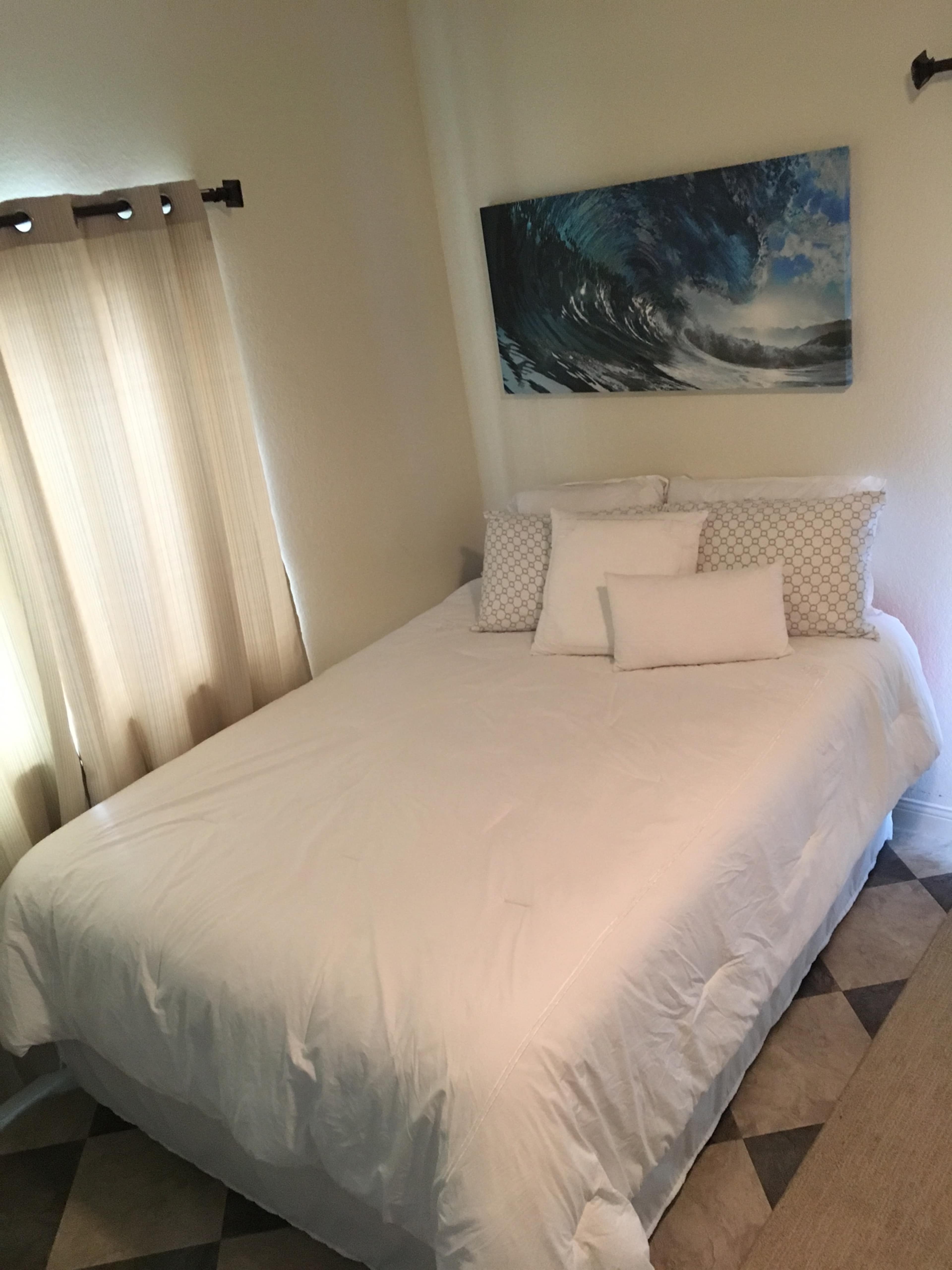 A bed with white bedding is positioned near a window with light-colored curtains, and a wave painting hangs on the wall above it.