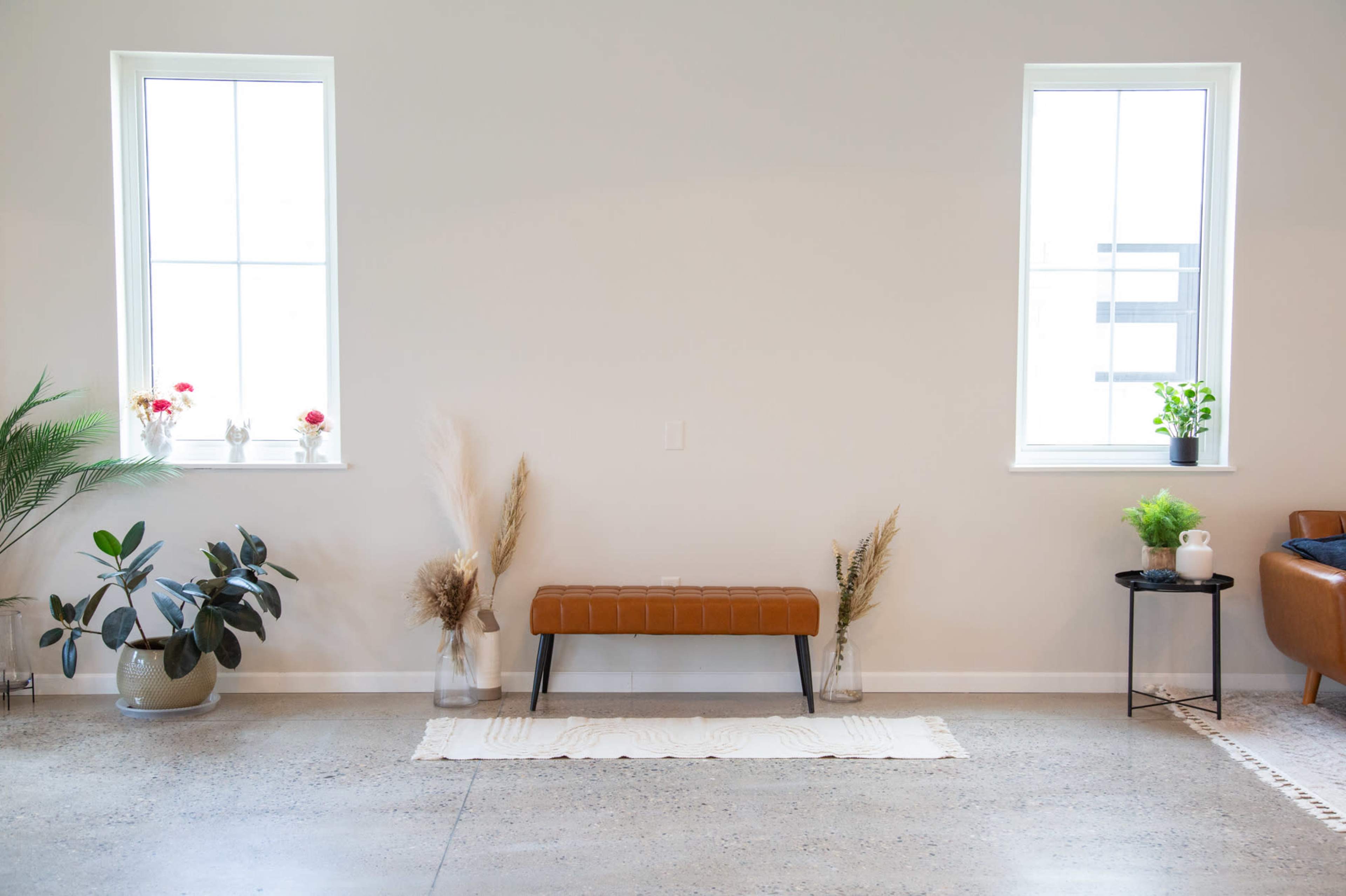A minimalist interior with a brown bench centered against a light-colored wall, flanked by plants and decorative elements near two windows.