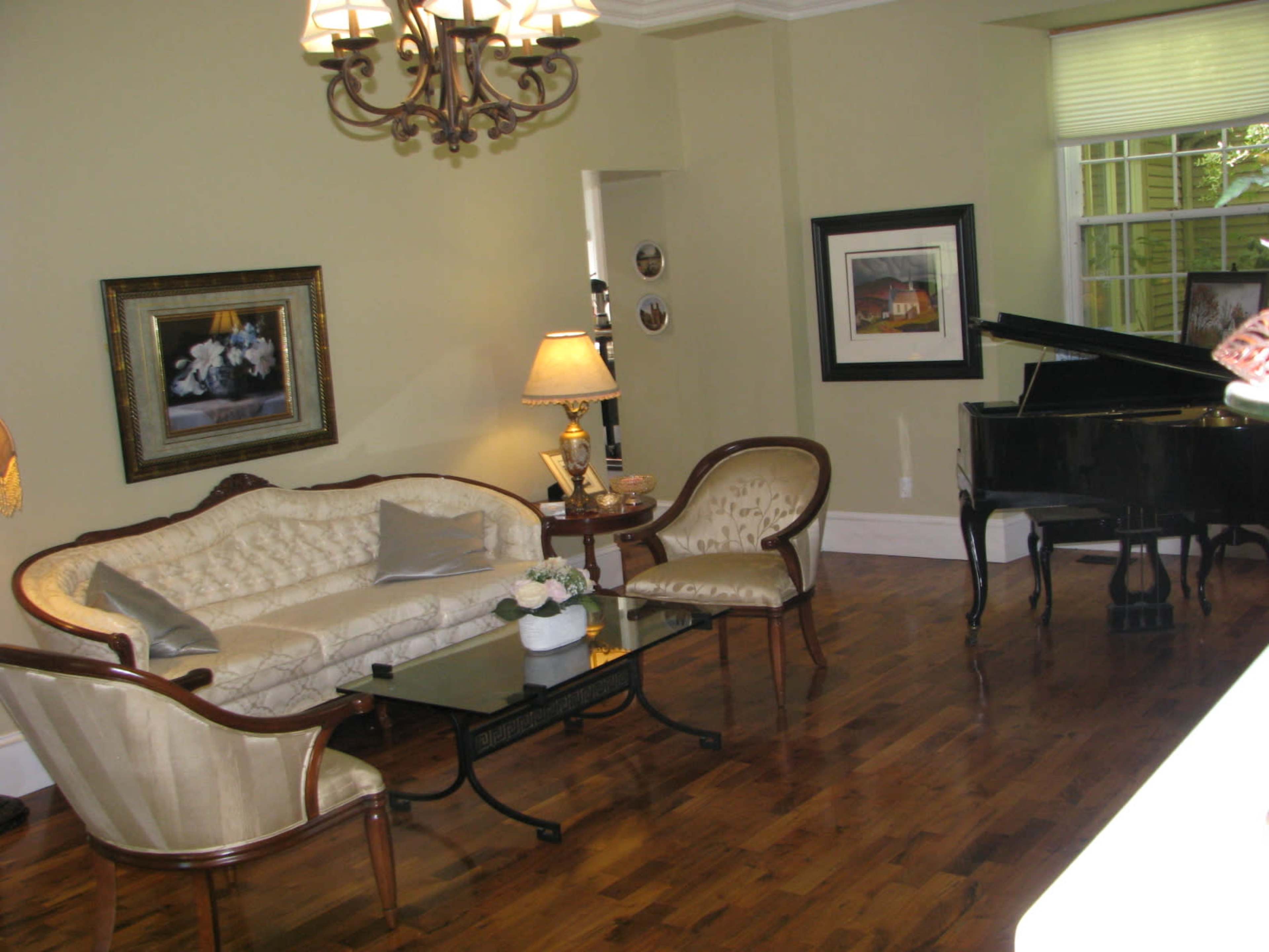 A living room features a cream-colored sofa, two elegant chairs, a glass coffee table, and a grand piano, all set against wooden flooring and light green walls.