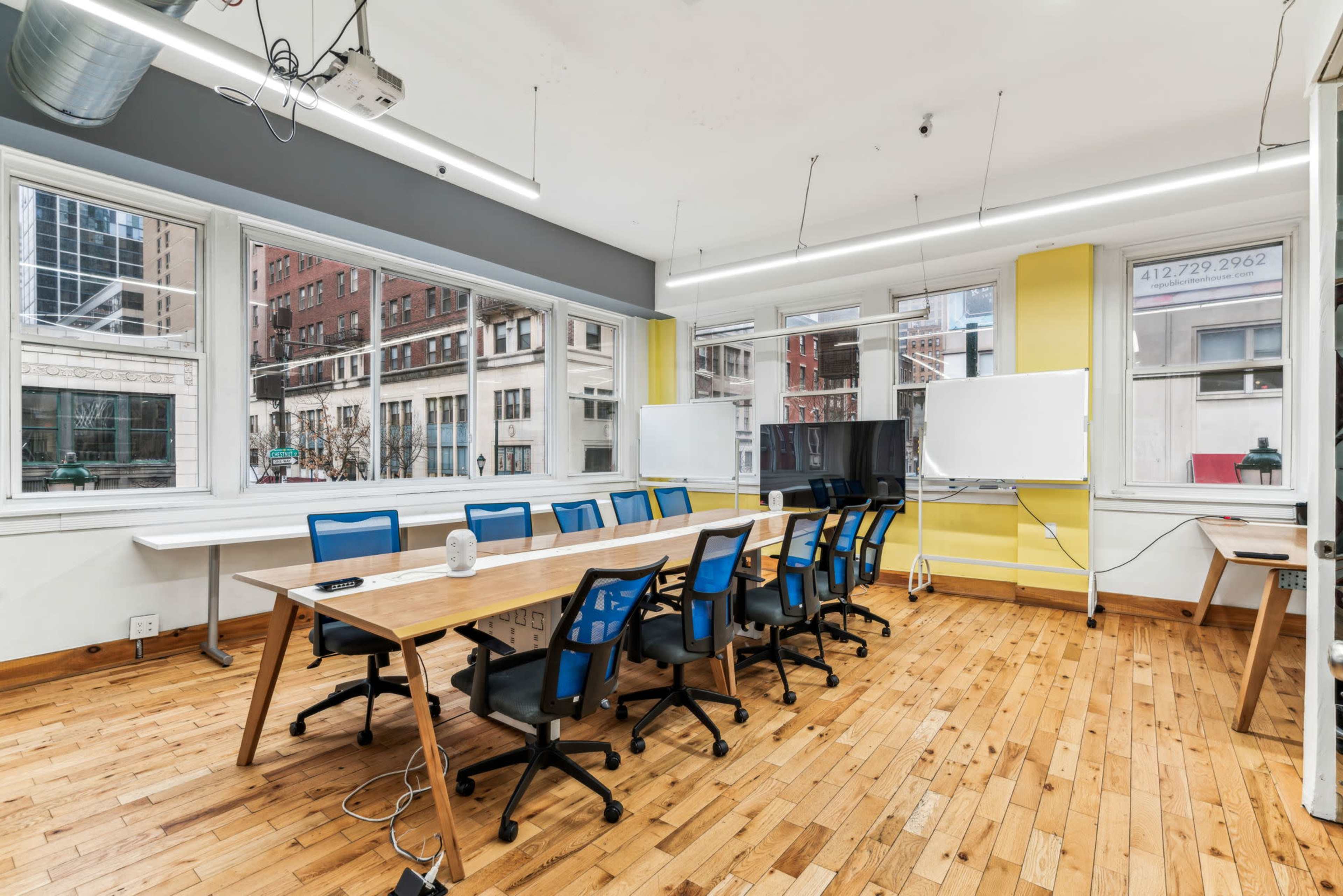A bright conference room features a long wooden table surrounded by blue office chairs, large windows, and whiteboards on the walls.