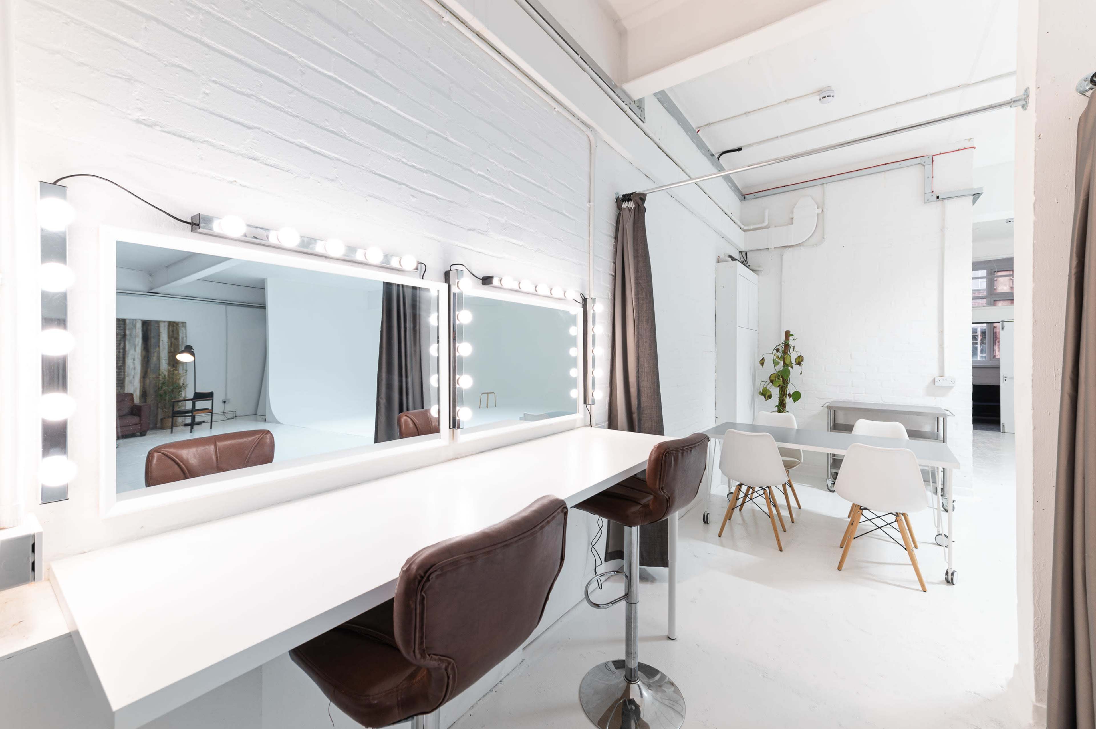 A well-lit studio space with two large mirrors surrounded by light bulbs, a white counter with brown stools, and tables and chairs in a separate area.