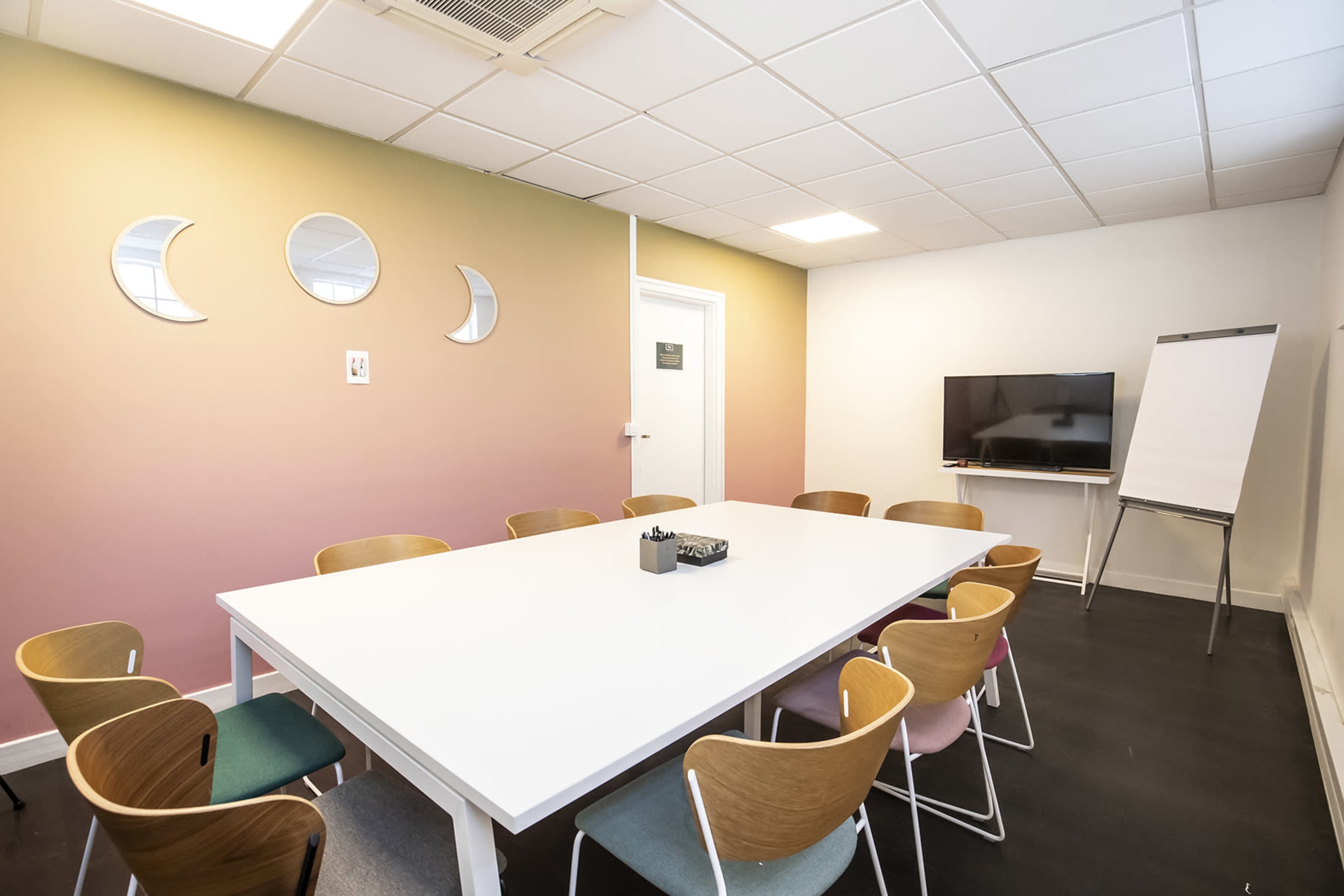 A meeting room featuring a large white table surrounded by wooden chairs, with a television and a flip chart at one end, and three circular mirrors on the wall.