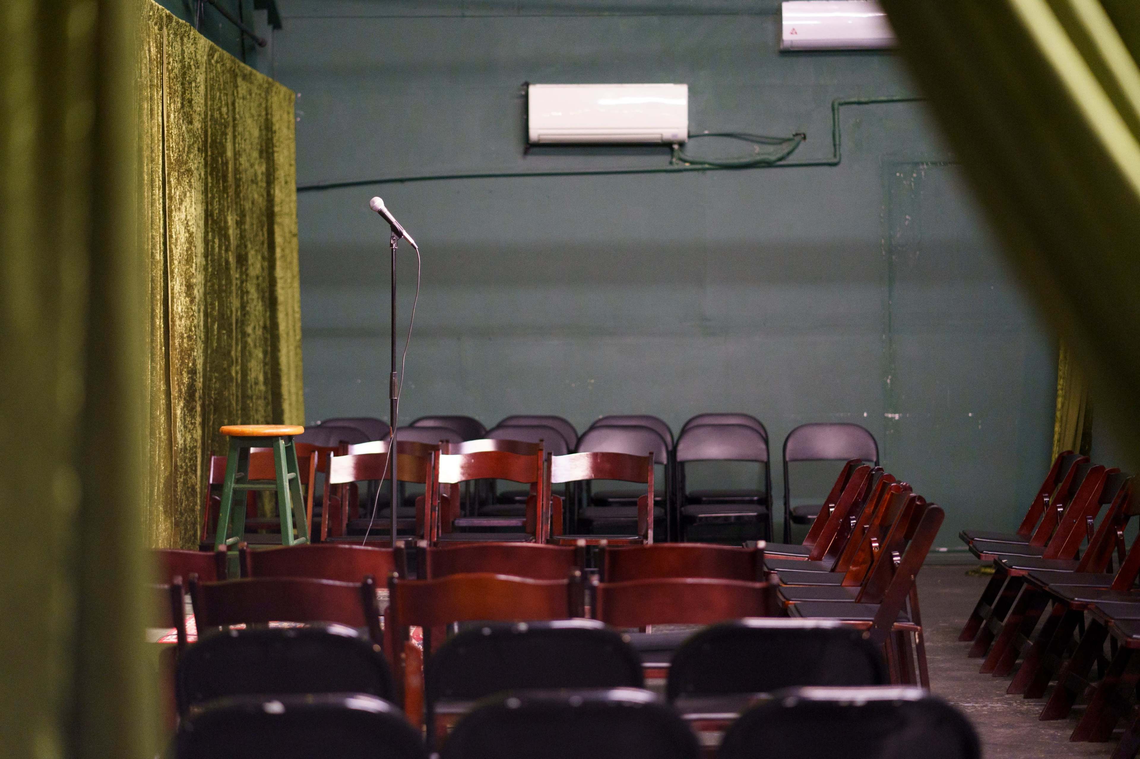 The image shows an empty comedy stage setup with a microphone on a stand and rows of chairs arranged in a dimly lit venue.